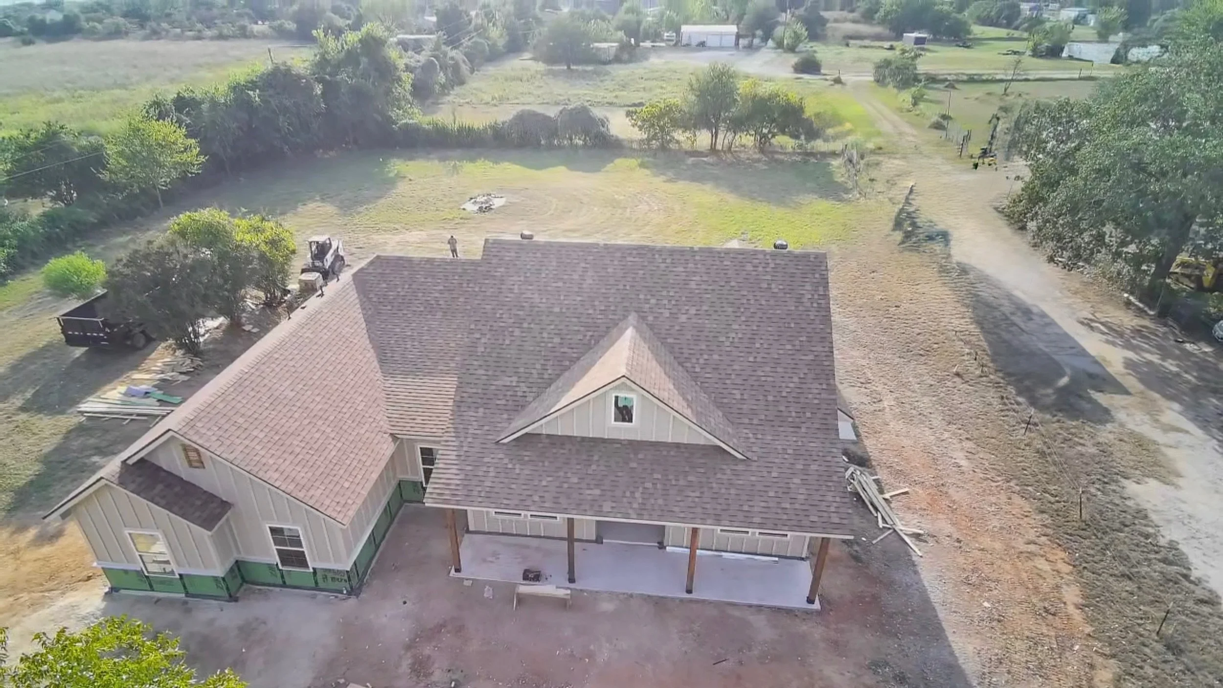An aerial view of a house under construction with parts of the exterior siding visible, a covered porch, a brown shingled roof, and a backyard. Construction materials and tools are scattered around, with construction workers and equipment present, and a dirt driveway leading to the house in a rural or semi-rural setting with trees and open fields surrounding the property.