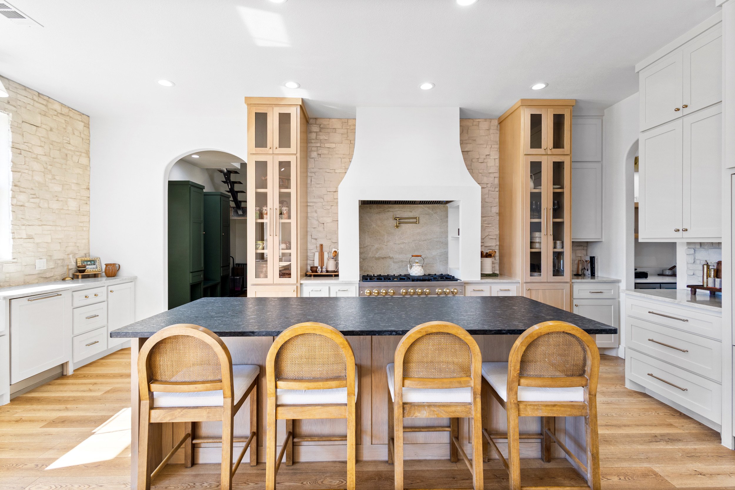 Modern kitchen with white cabinets, a black countertop island, and a gas range in the center with a stone backsplash. Wooden bar stools are in front of the island, and the room has hardwood flooring and recessed ceiling lights.
