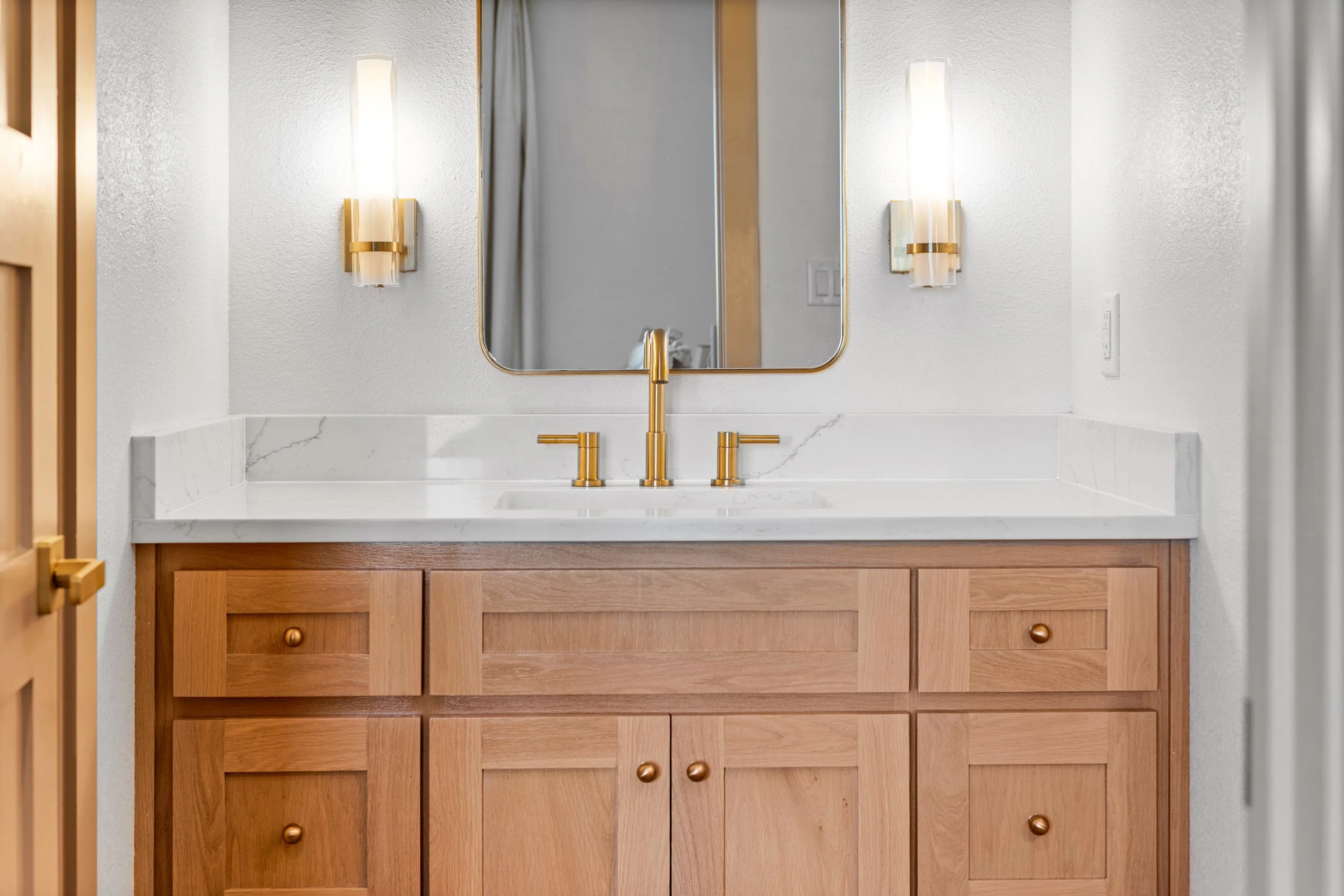 Bathroom vanity with a wooden cabinet, white quartz countertop, brass faucet, rectangular mirror, and two wall-mounted light fixtures.