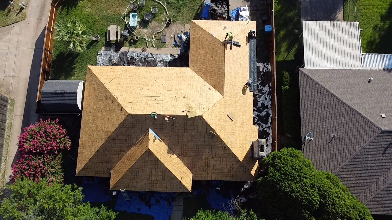 An aerial view of a house with a yellowish brown roof, surrounded by a fenced backyard. There are workers on the roof, and construction materials scattered around. The backyard has trees, including a flowering pink tree and green leafy trees, as well as outdoor furniture and gardening tools.