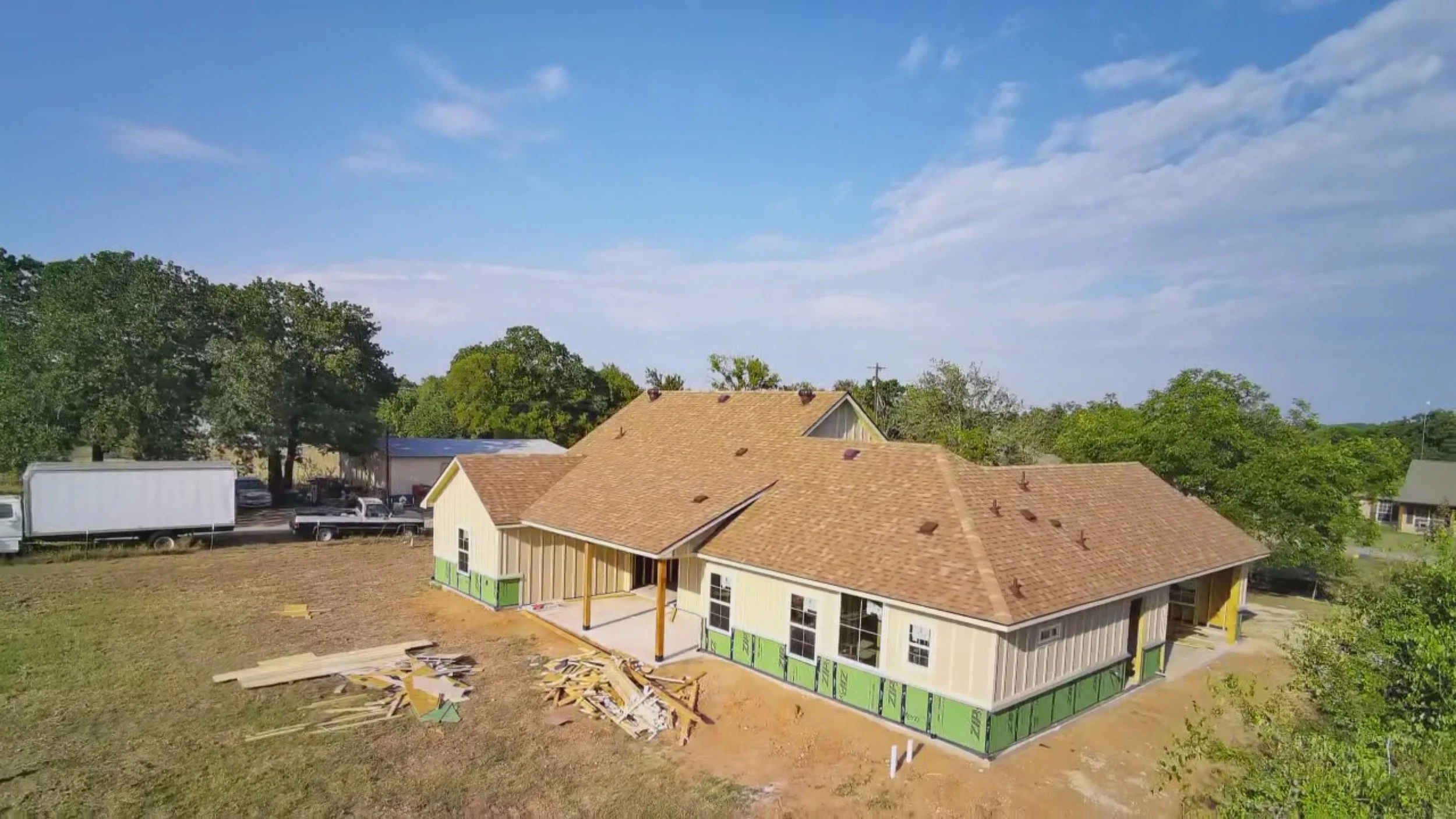 A house under construction with a tan roof and cream walls, surrounded by green trees and equipment, on a clear day with a partly cloudy sky.