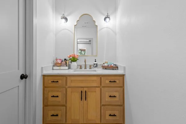 Bathroom vanity with a wooden cabinet, white countertop, soap dispenser, and a pink flower arrangement, reflected in an arched mirror surrounded by two wall-mounted lights, with white walls.
