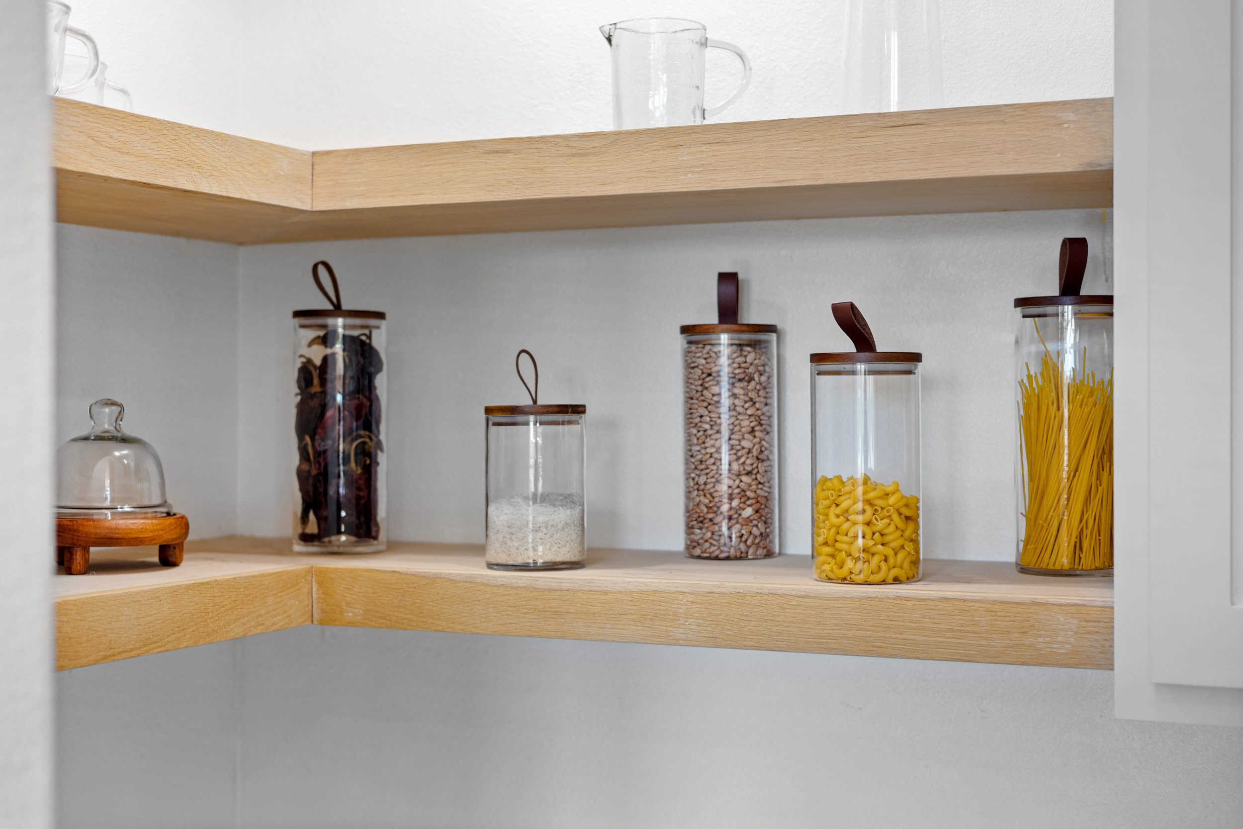 Kitchen pantry shelf holding glass jars filled with dried pasta, beans, flour, and dried herbs, with a white wall background.