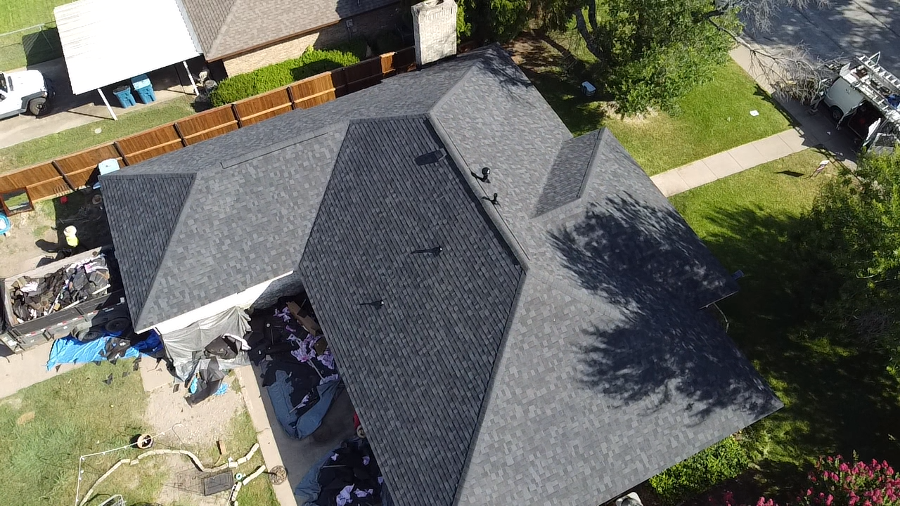 Aerial view of a house with a dark gray shingle roof and a backyard cluttered with various objects and debris, with a green lawn and trees surrounding the property.