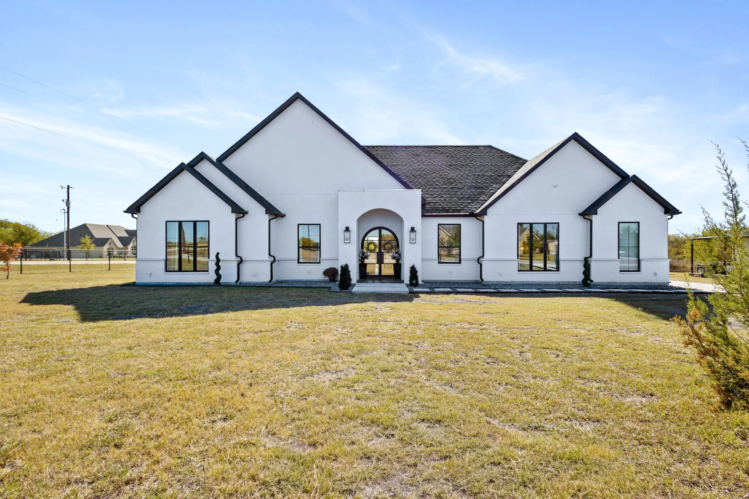 Front view of a modern white house with black roof and window frames, surrounded by a grassy yard on a sunny day.