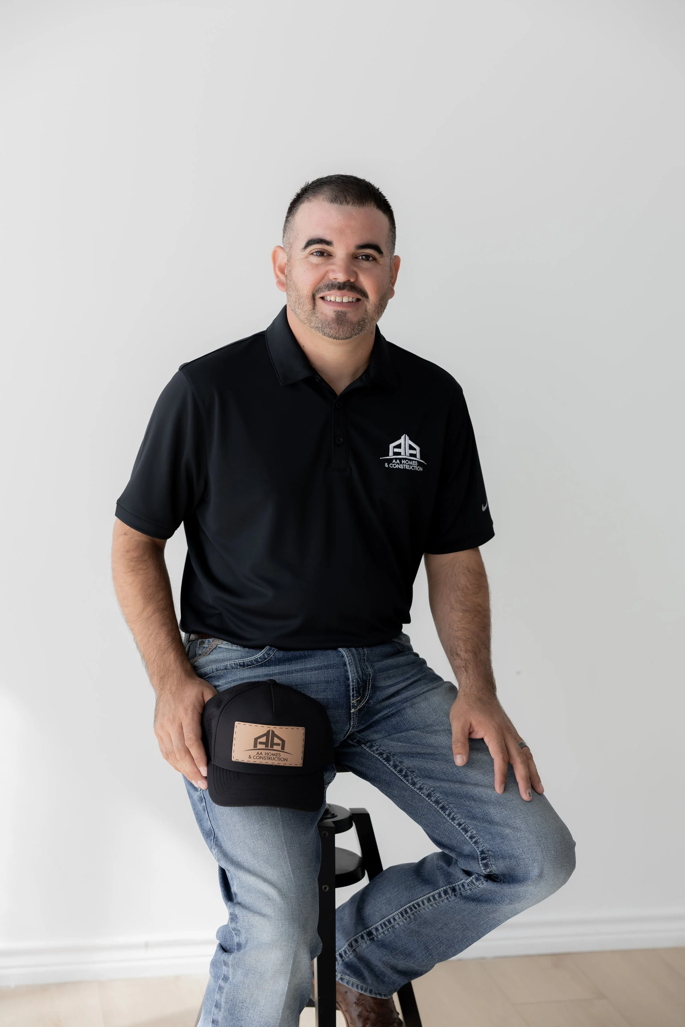 A man with short dark hair and a beard sits on a stool, wearing a black polo shirt with a construction company logo and holding a black cap with the same logo against a plain white background.