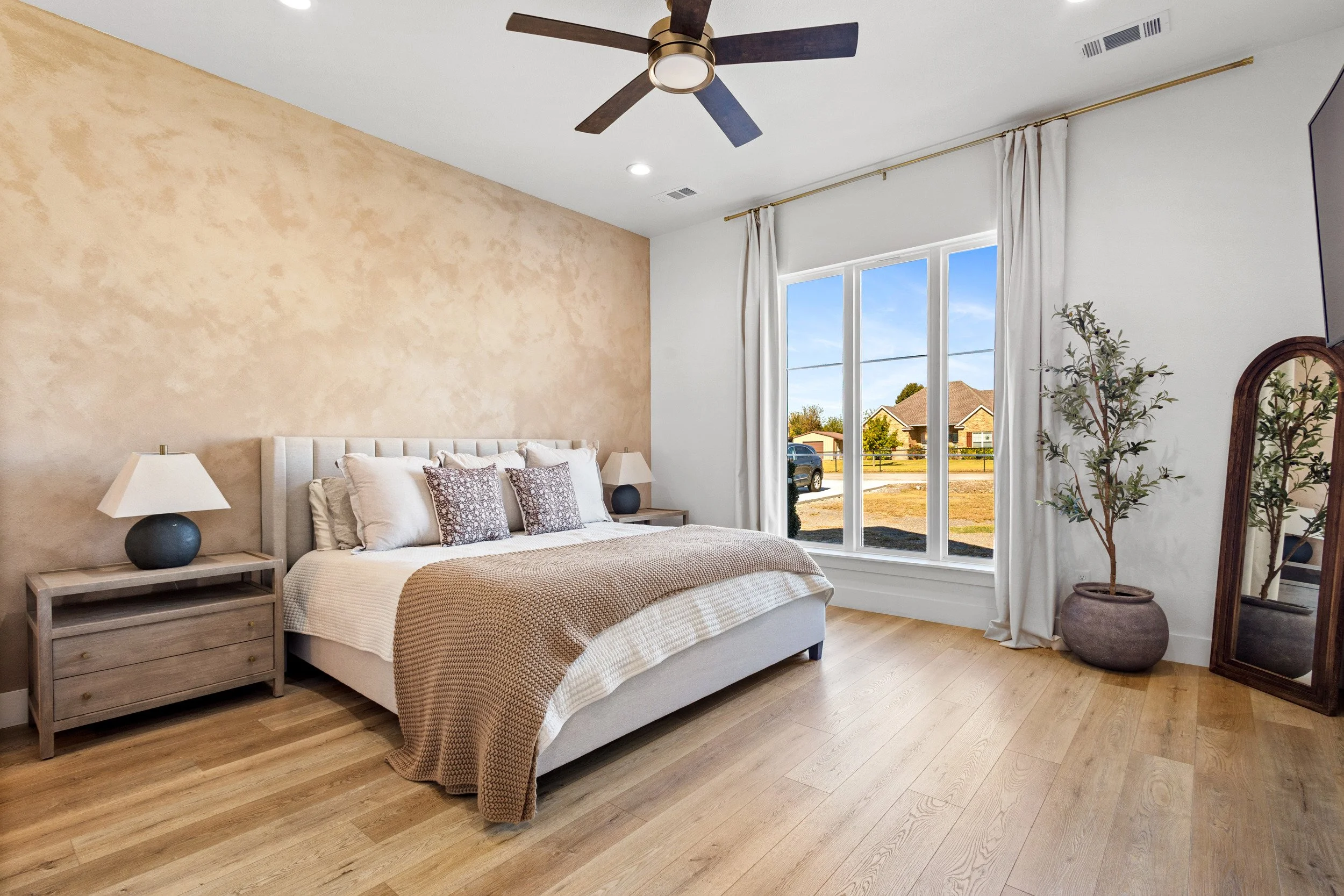 A bedroom with a large window showing a rural neighborhood, a bed with beige and white bedding, two nightstands with lamps, a ceiling fan, a tall mirror, and a potted plant.
