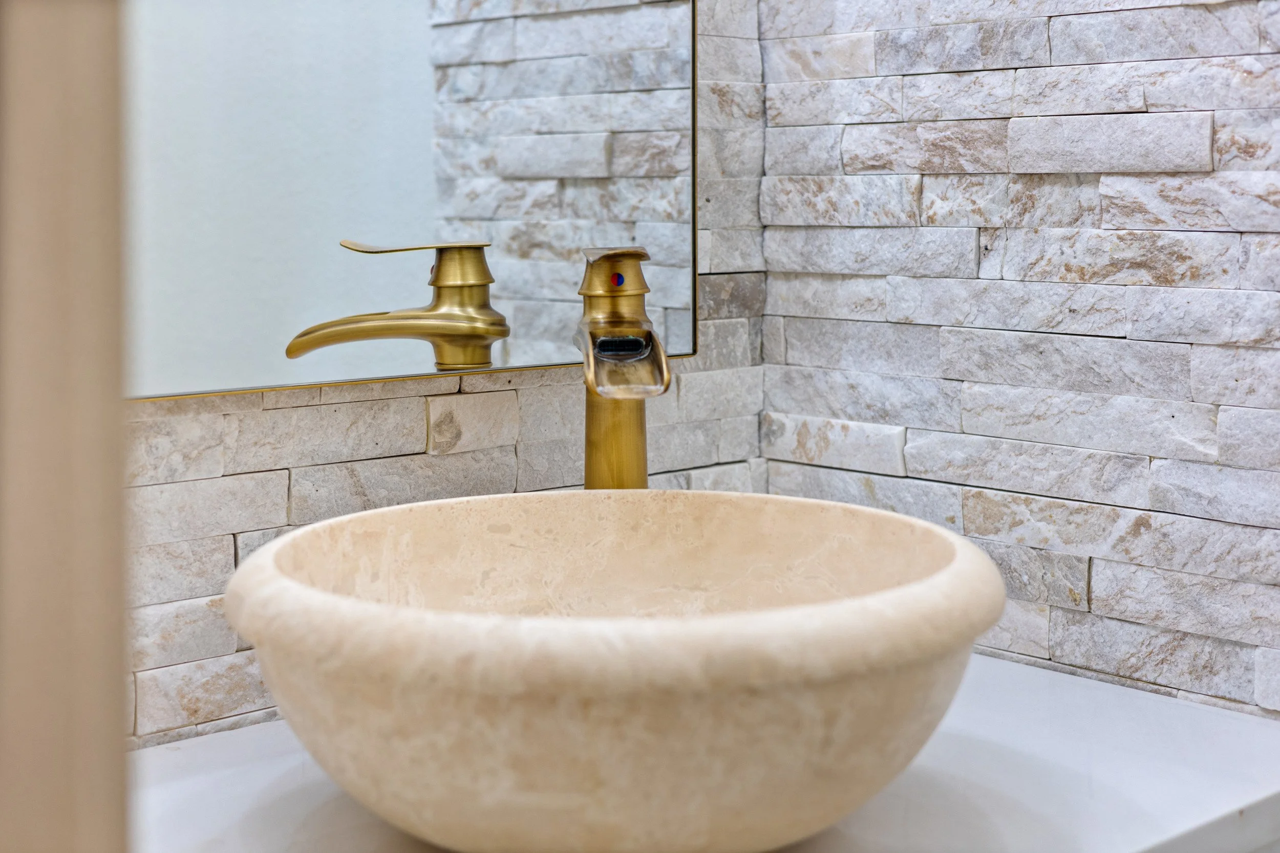 A bathroom sink with a beige stone bowl, a gold faucet, and a mirror reflecting a textured stone tile wall.