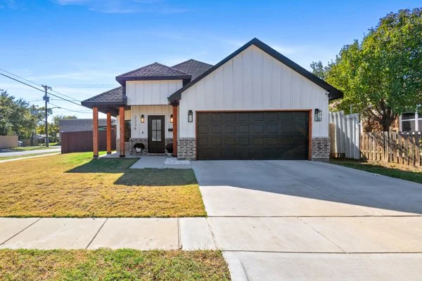 A modern two-story house with a dark garage door, white siding, brick accents, and a small front porch, surrounded by a lawn and a concrete driveway.