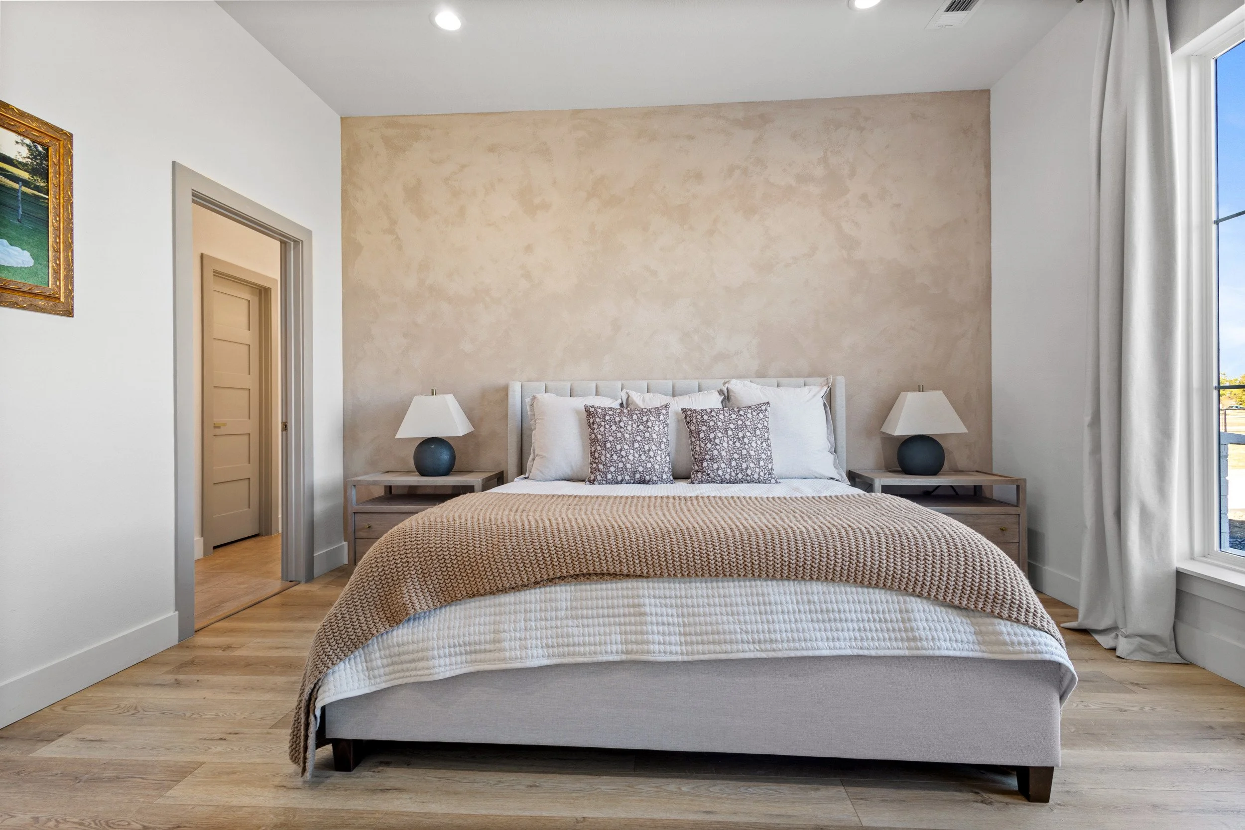 Well-made bedroom with a bed, beige blanket, and two nightstands with lamps, large window with curtains, and a door leading to another room.