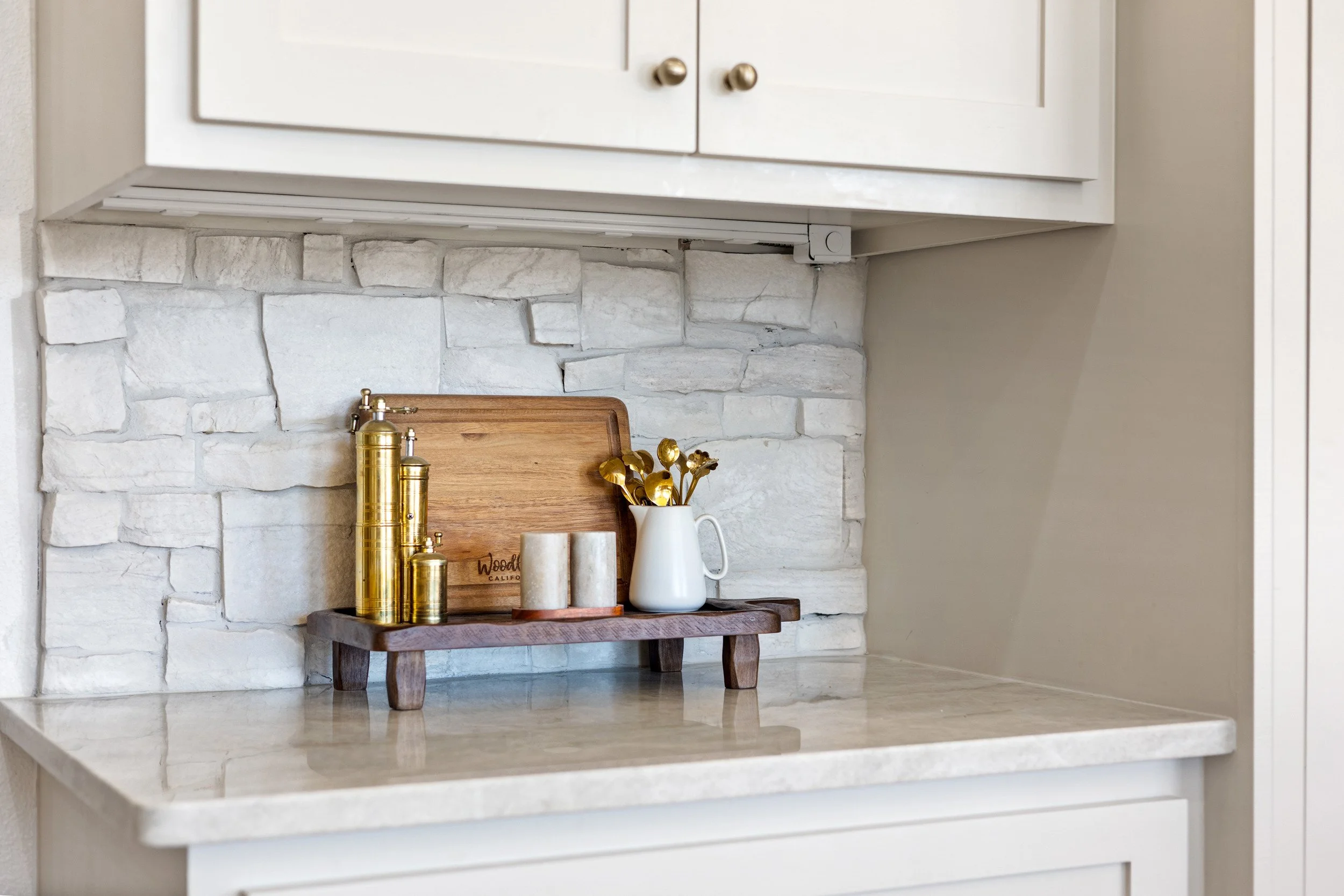 Decorative items on a small wooden tray, including a tall gold salt or pepper shaker, two smaller gold containers, a white ceramic vase with gold spoons, and a wooden cutting board, arranged against a white stone wall in a kitchen.