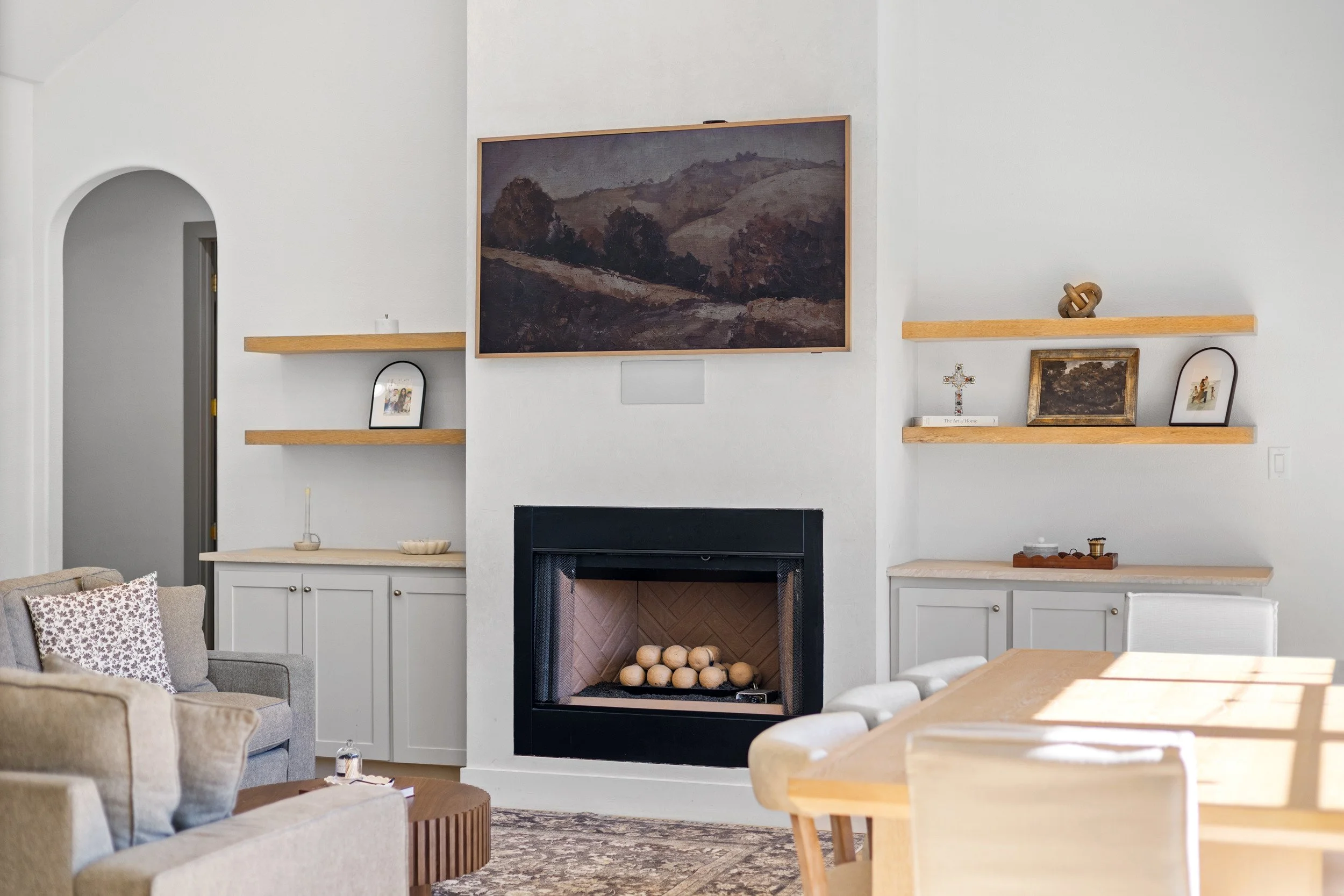 Living room with white walls, a fireplace, and wall-mounted wooden shelves holding framed pictures, a cross, and decorative items. A large landscape painting hangs above the fireplace, and there is a seating area with a gray sofa, beige armchair, and