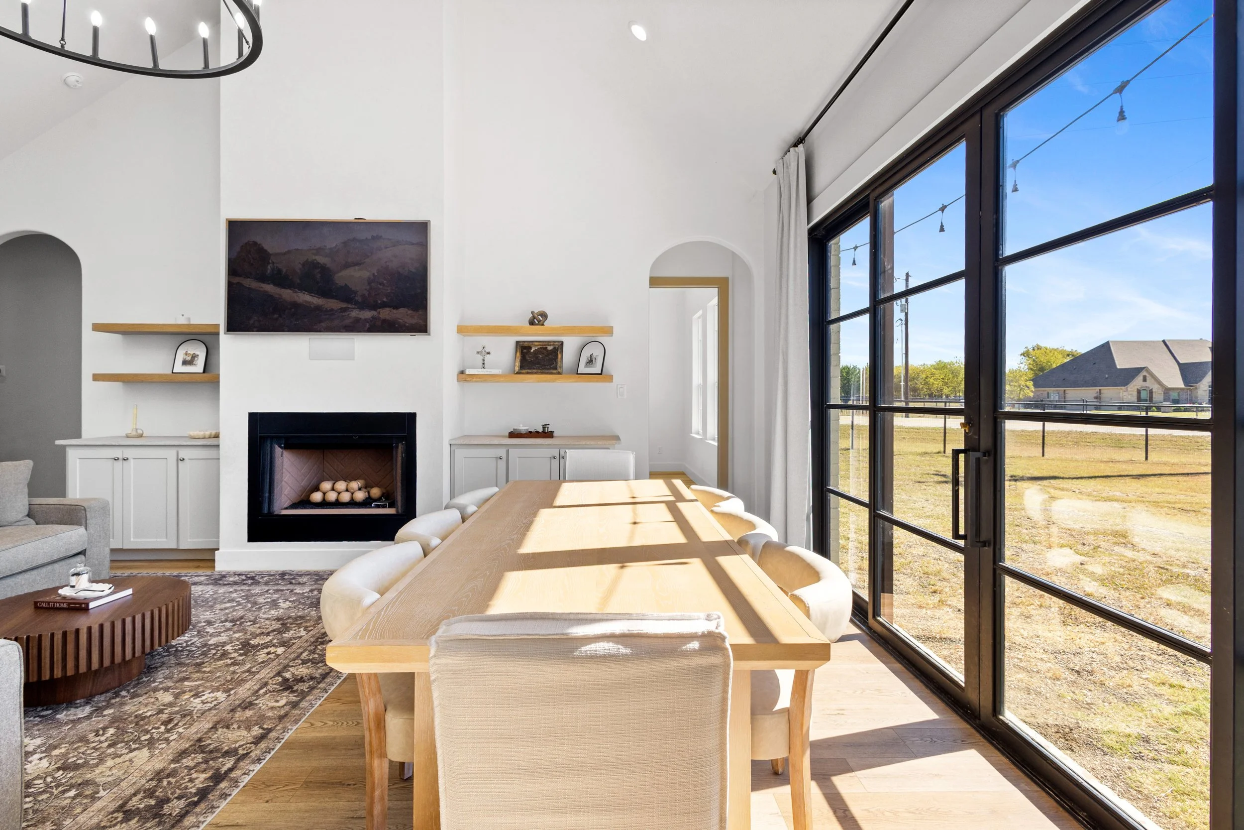 Interior of a modern dining room with a large wooden table, beige upholstered chairs, a white fireplace, and a view of the outdoors through large glass sliding doors.