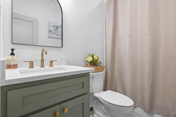 Bathroom with a green vanity, gold faucet, mirror, soap dispenser, flower arrangement, and beige shower curtain.