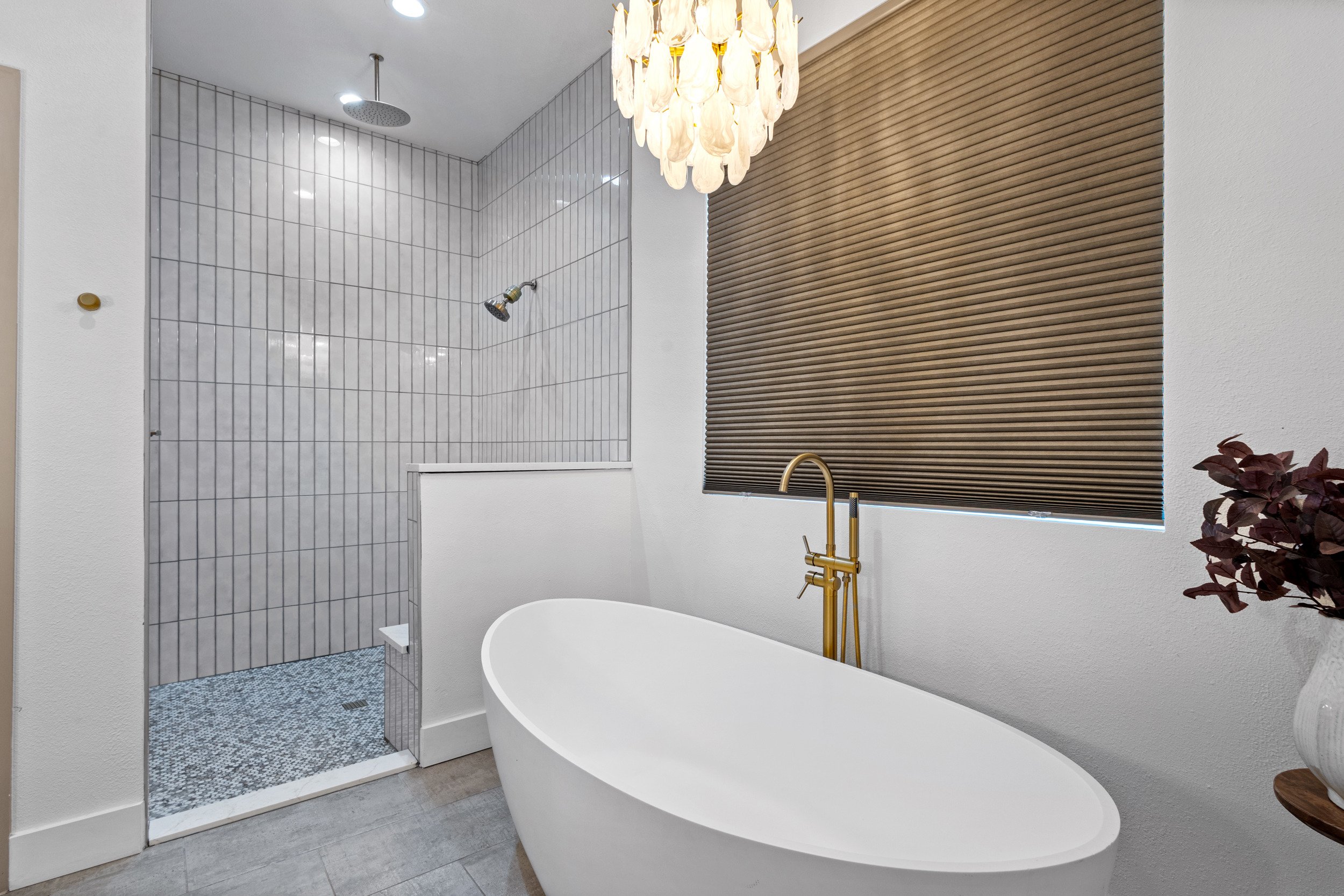 Modern bathroom with a white freestanding bathtub, white tiled shower area with a rainfall showerhead, brown window blind, and a decorative plant in a vase.