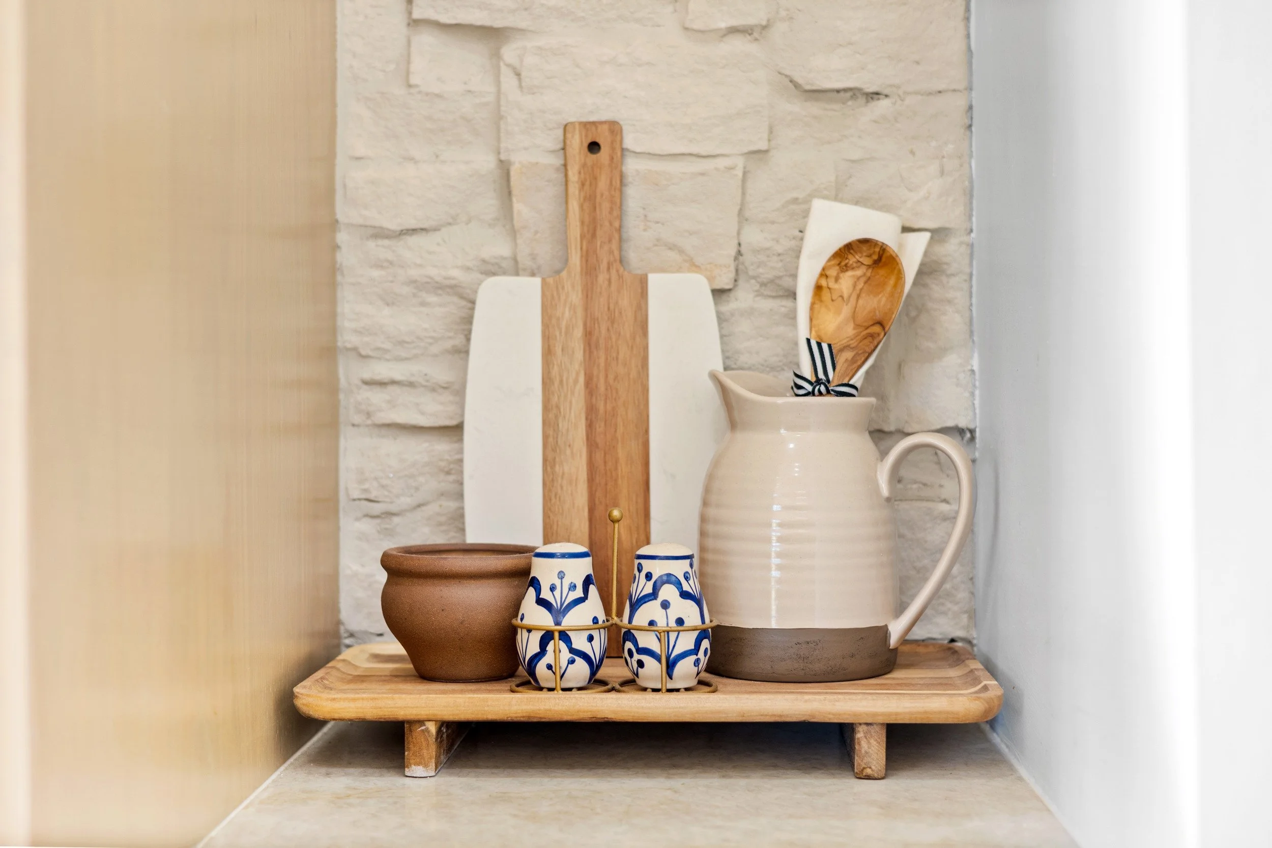 Kitchen countertop display with a wooden tray holding a small brown bowl, a pair of blue and white patterned salt and pepper shakers, a beige ceramic pitcher with a handle, a wooden spoon inside the pitcher, and a white spatula with a black ribbon, a