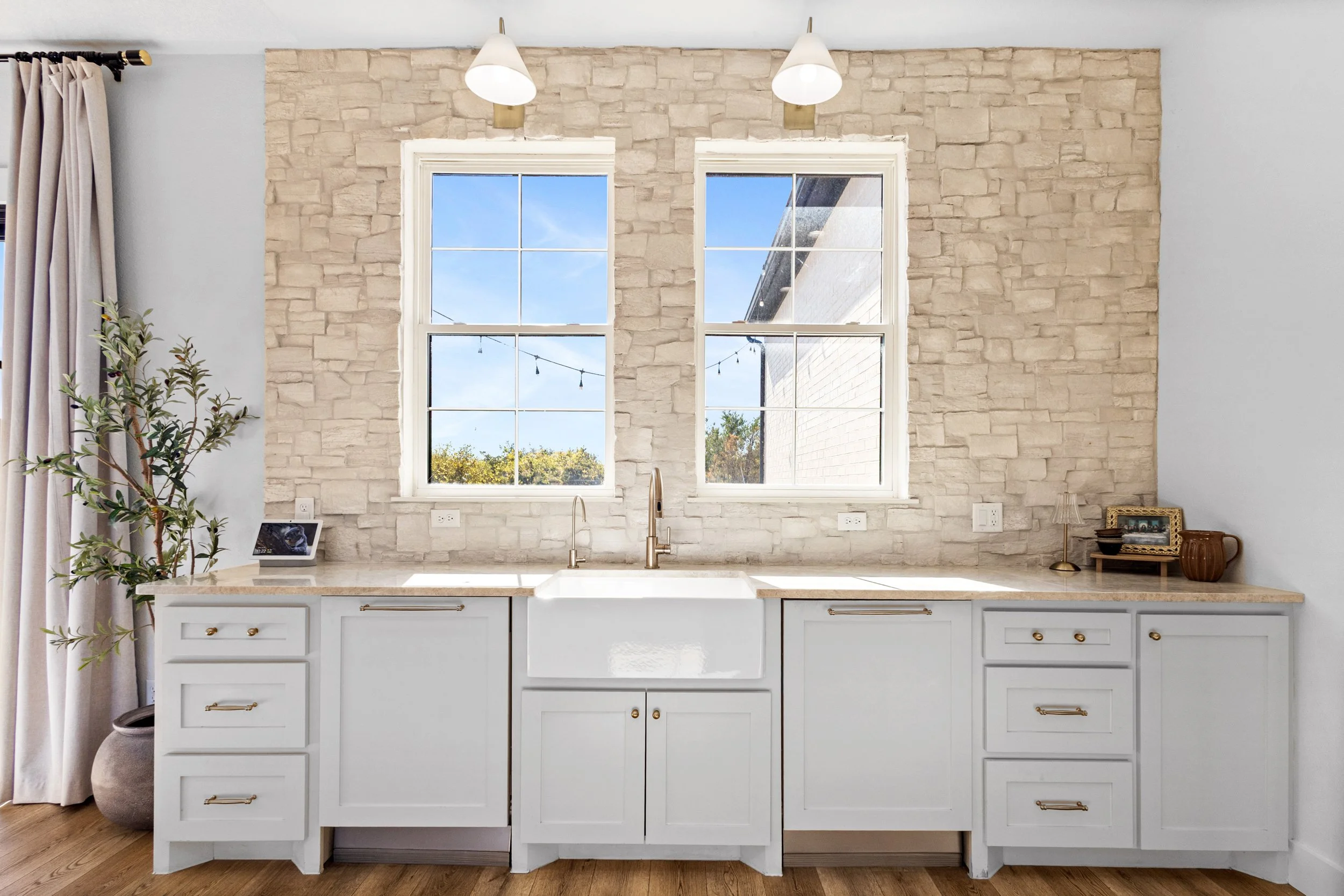 Kitchen with white cabinetry, farmhouse sink, two windows with views of a blue sky and trees, stone wall backdrop, and pendant lights extending from the wall.