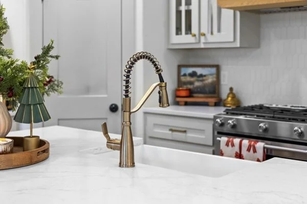 Close-up of a kitchen sink with a gold-colored faucet on a white quartz countertop, with a Christmas tree decoration on a wooden tray to the side.