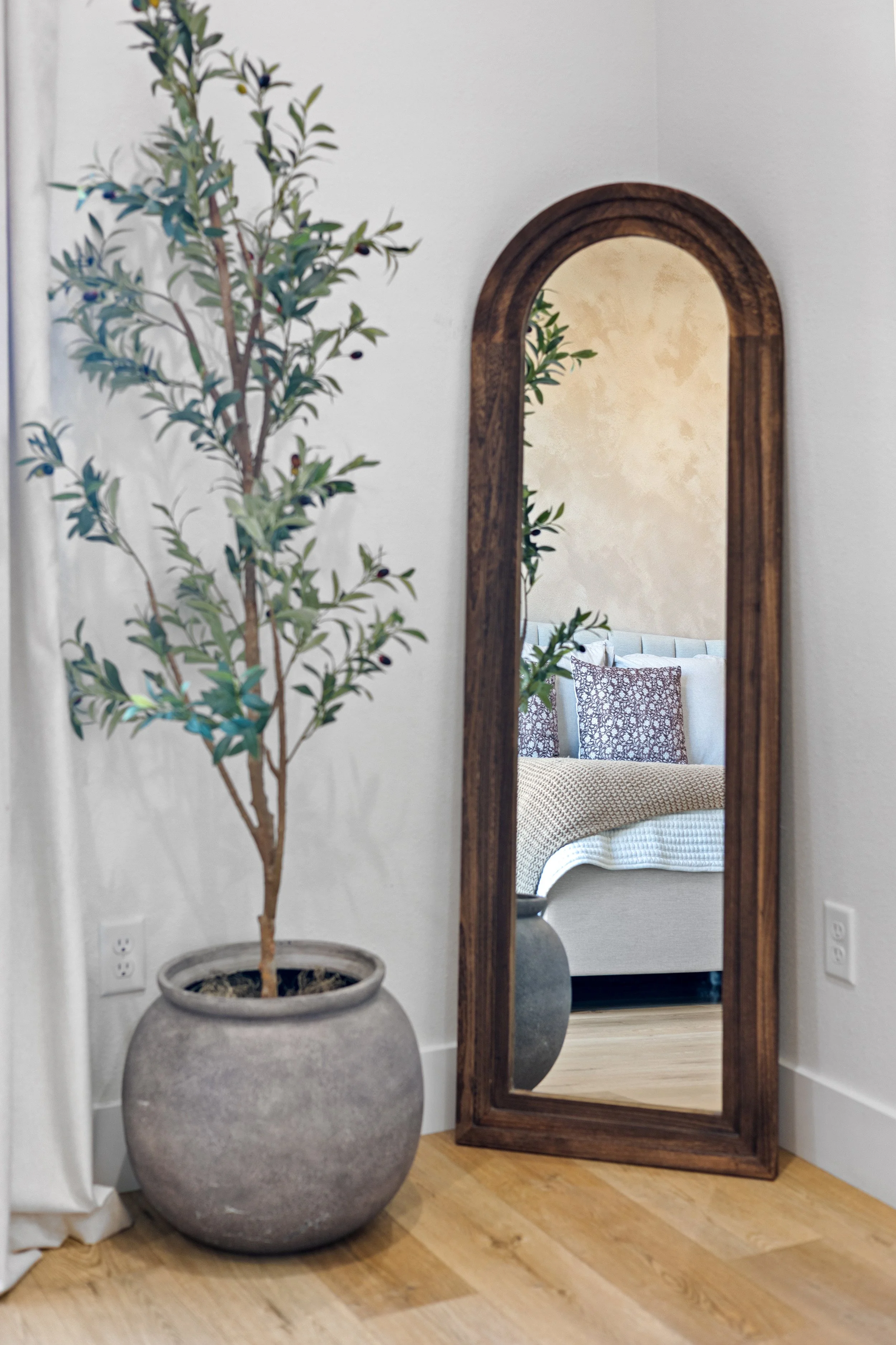 A potted plant with green leaves and small dark berries next to a tall wooden-framed mirror reflecting a sofa with patterned pillows.