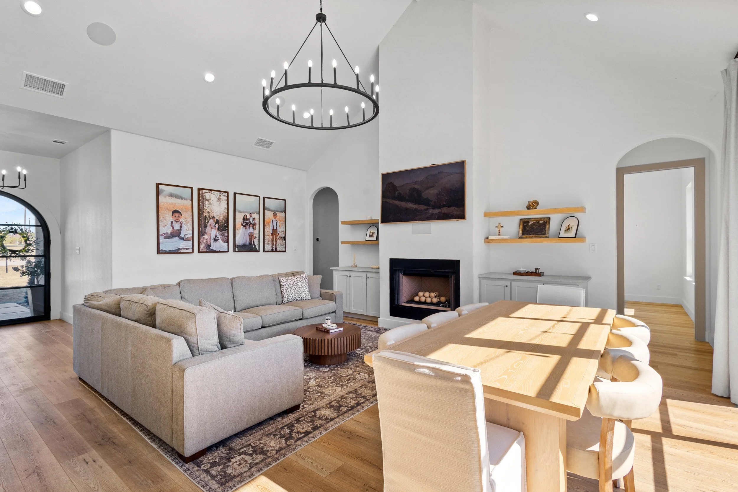 Bright living room with white walls, wooden flooring, and modern furniture. Contains a beige couch, a wooden coffee table, a dining table with white chairs, and a black-framed fireplace. Decorated with framed photos and paintings, and a large chandel