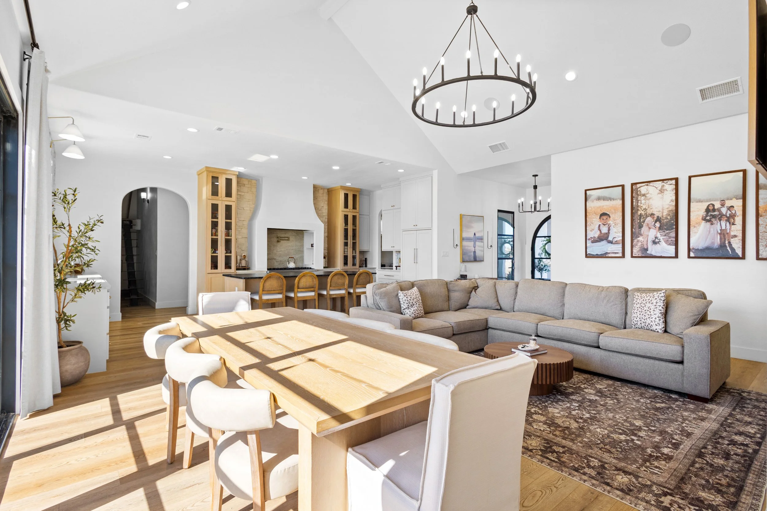 A spacious, modern open-concept living room and kitchen with hardwood floors and white walls. The living area features a large beige sectional sofa, a patterned area rug, and a wooden coffee table. The dining area has a wooden table with six upholste