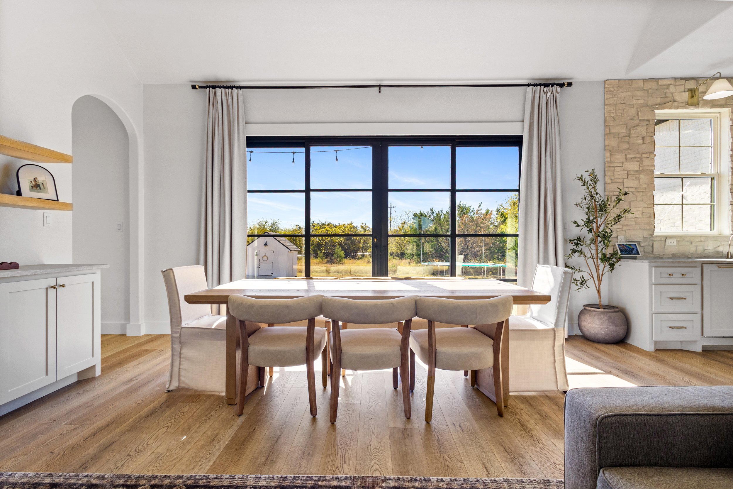 Bright dining room with a large window showing an outdoor view, surrounded by white walls and light wood flooring, featuring a wooden dining table with six chairs, a potted plant, and a white cabinetry area.