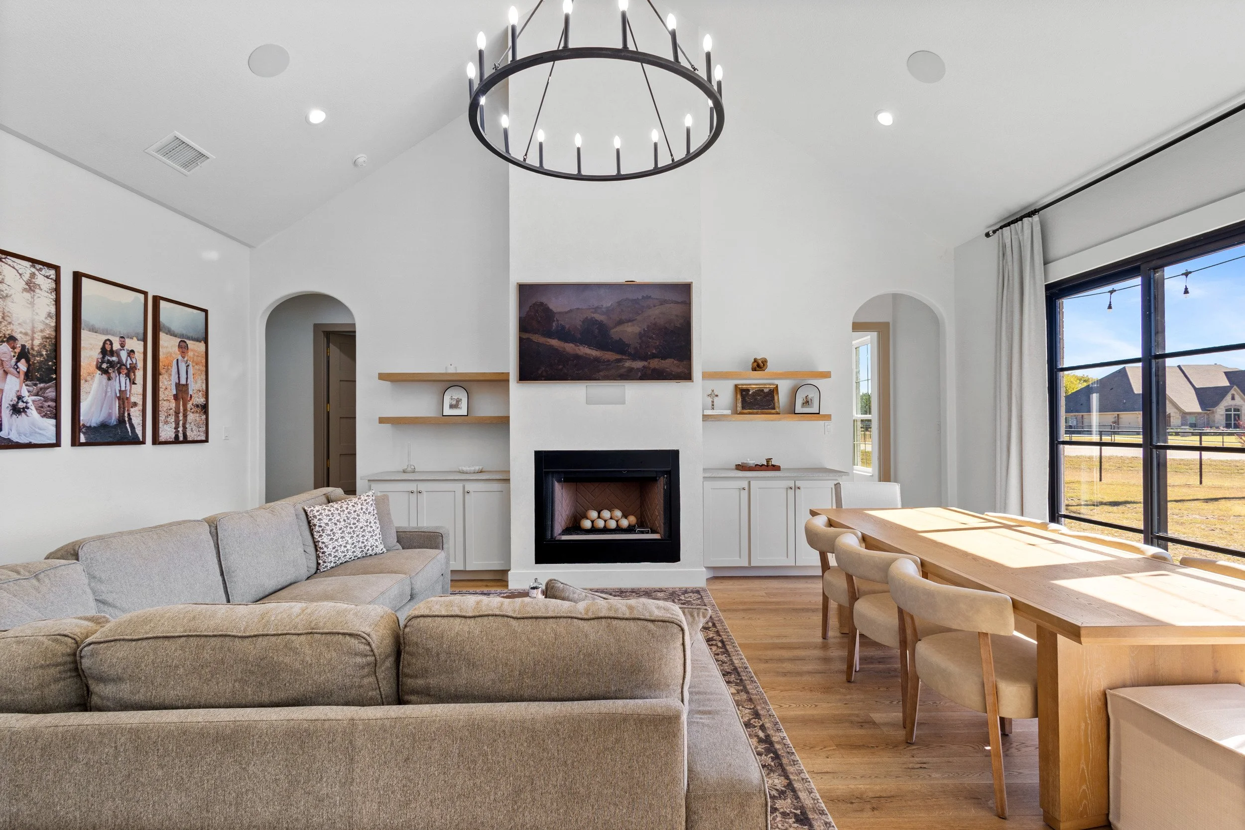 Bright living room with beige sectional sofa, white built-in cabinets, a wall-mounted TV above a fireplace with candles, a wooden dining table with beige chairs, large windows with white curtains, and decorative items on shelves.