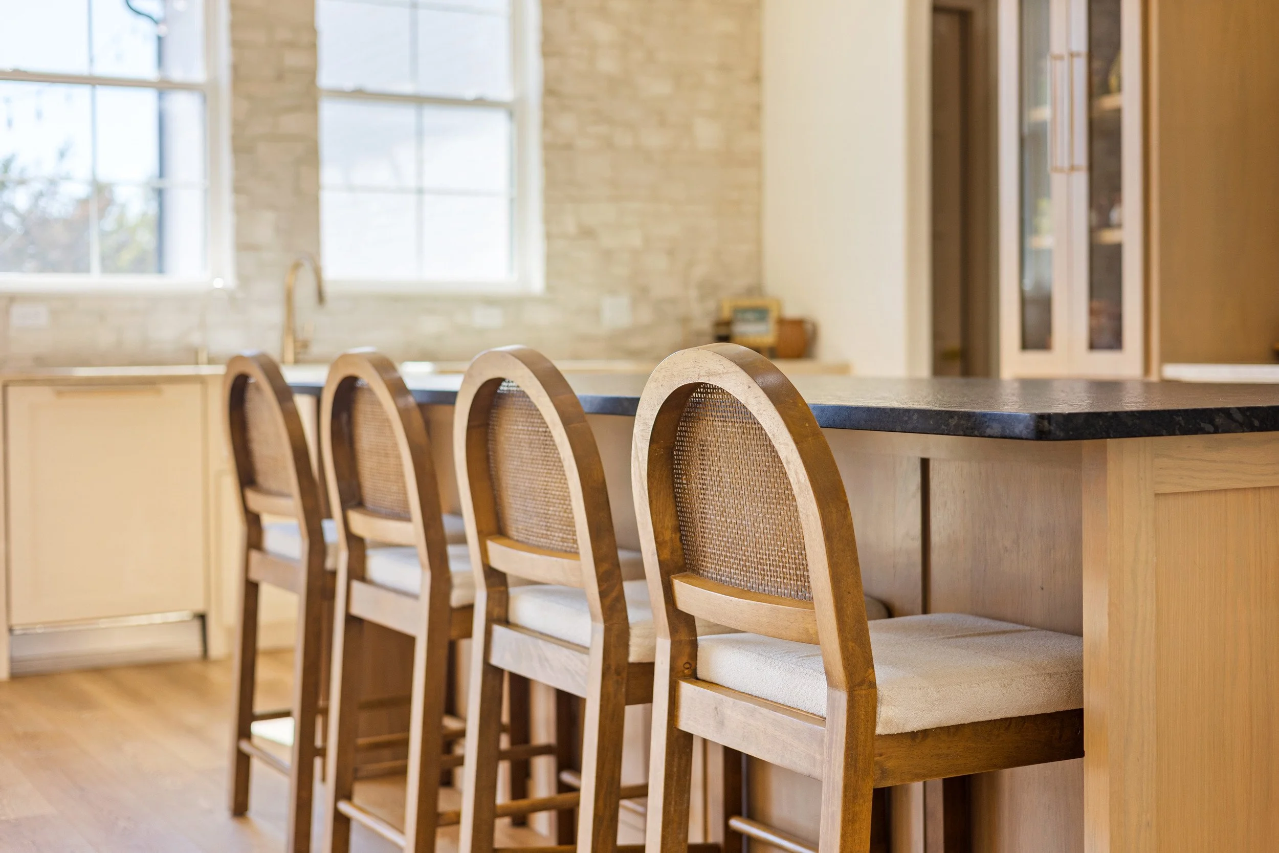A kitchen with a wooden island and four wooden barstools with cushioned seats, in front of a window with beige walls and cabinets.