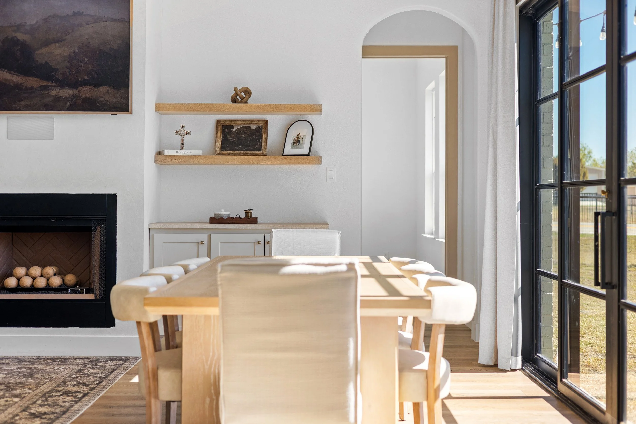 Dining room with a wooden table and cream-colored chairs near a large glass door; white walls with two wooden shelves holding framed pictures and decorative objects; a fireplace on the left; sunlight streaming in through the door.