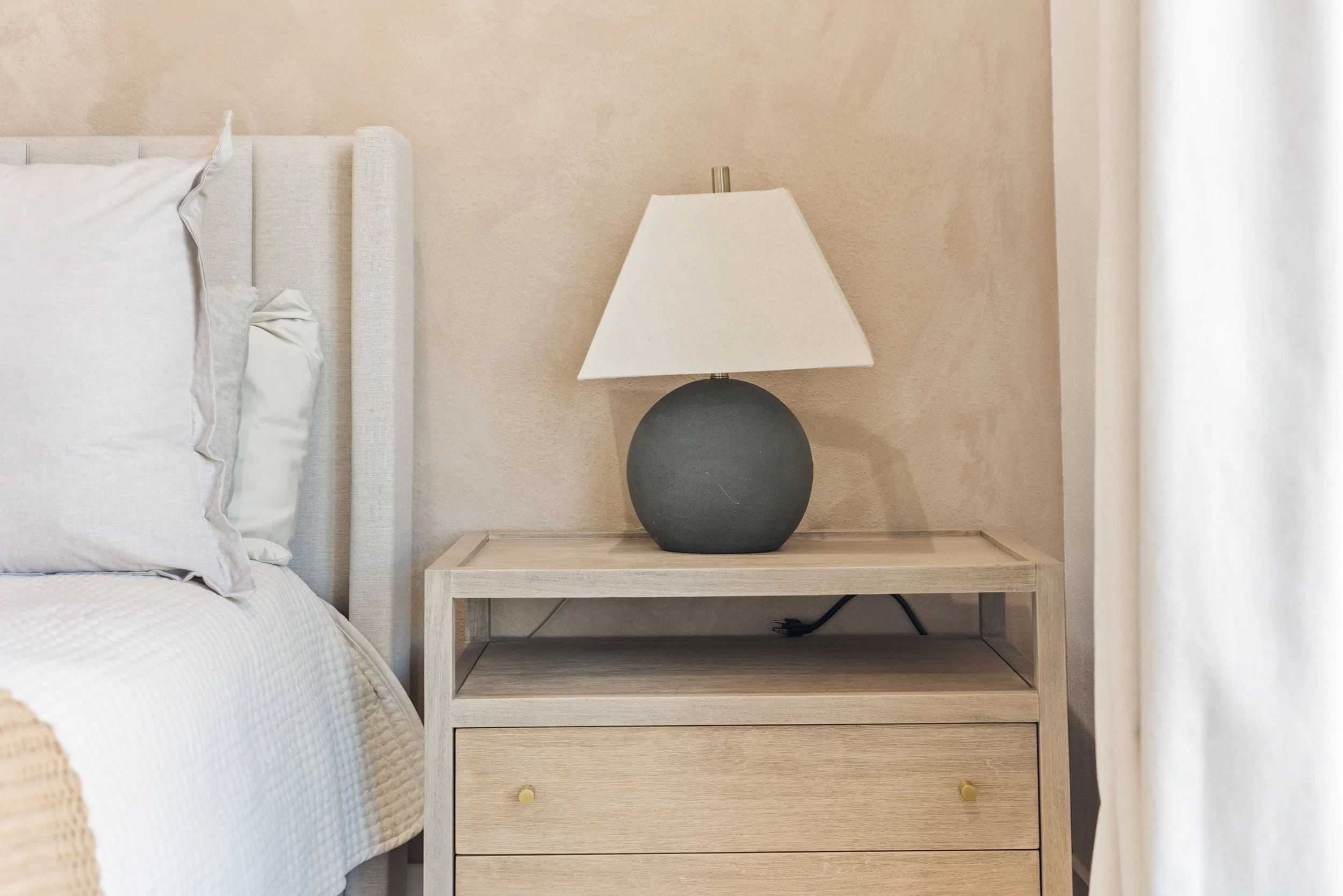 A bedroom nightstand with a lamp, next to a bed with white bedding and pillows, and a beige curtain on the right.