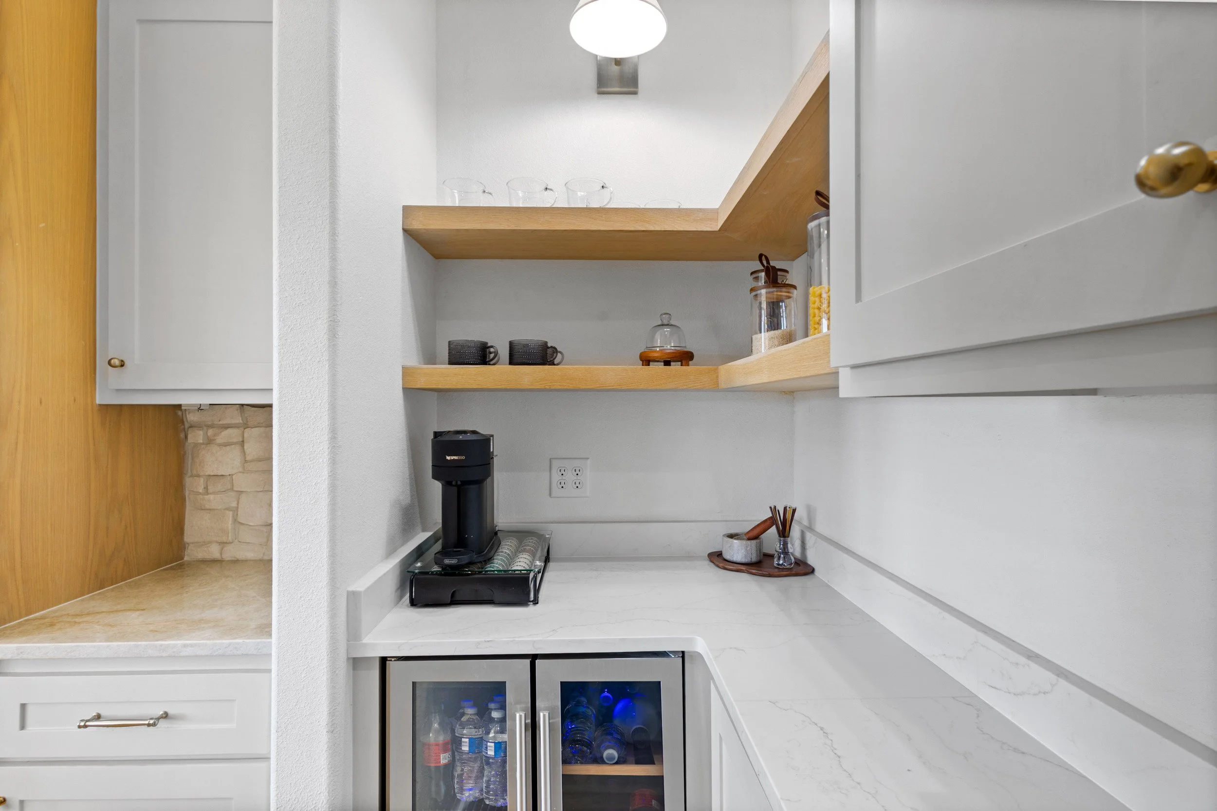 A modern kitchen corner with open wooden shelves, a coffee machine, water bottles in a mini fridge, and various kitchen items on the countertop.