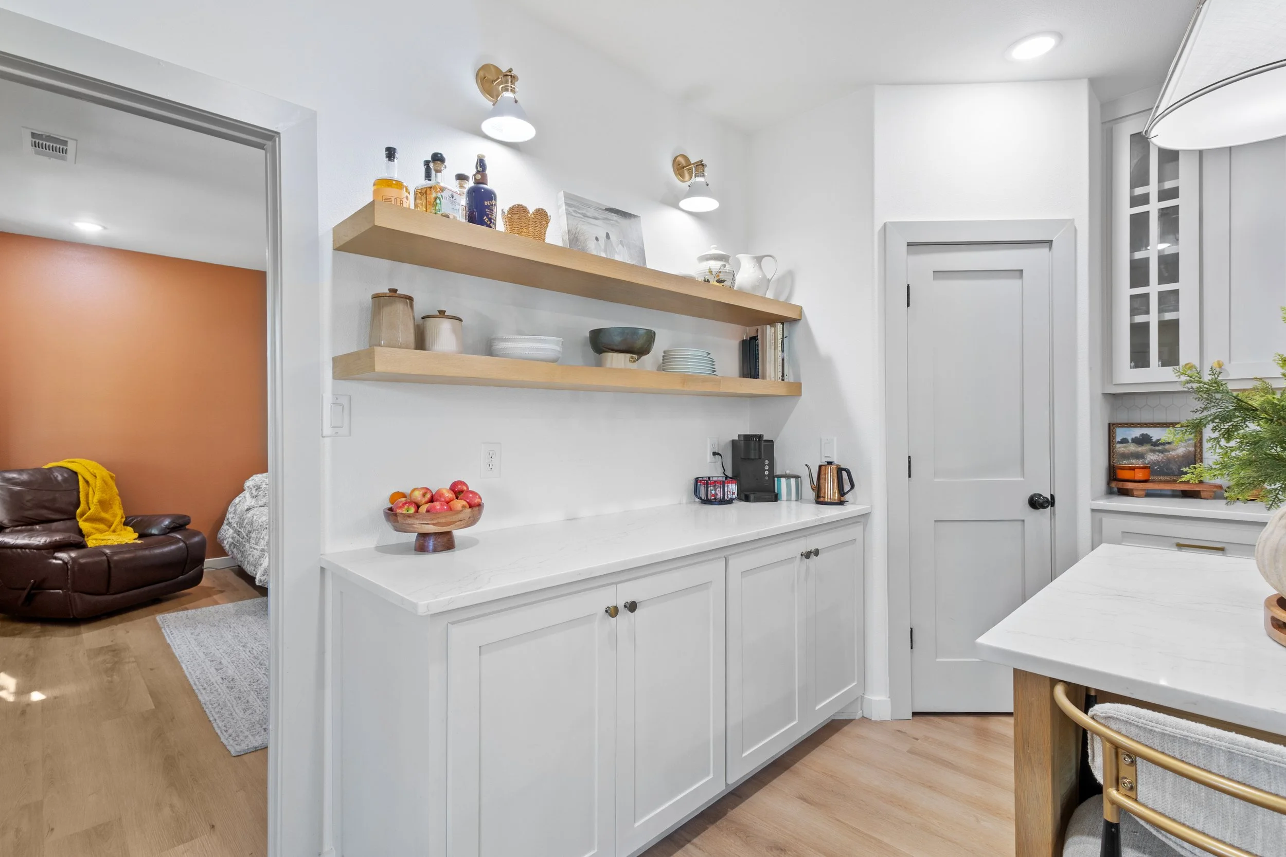 Modern kitchen with white cabinets, open wooden shelves, and a quartz countertop, with a doorway leading to a living room with a brown leather sofa.