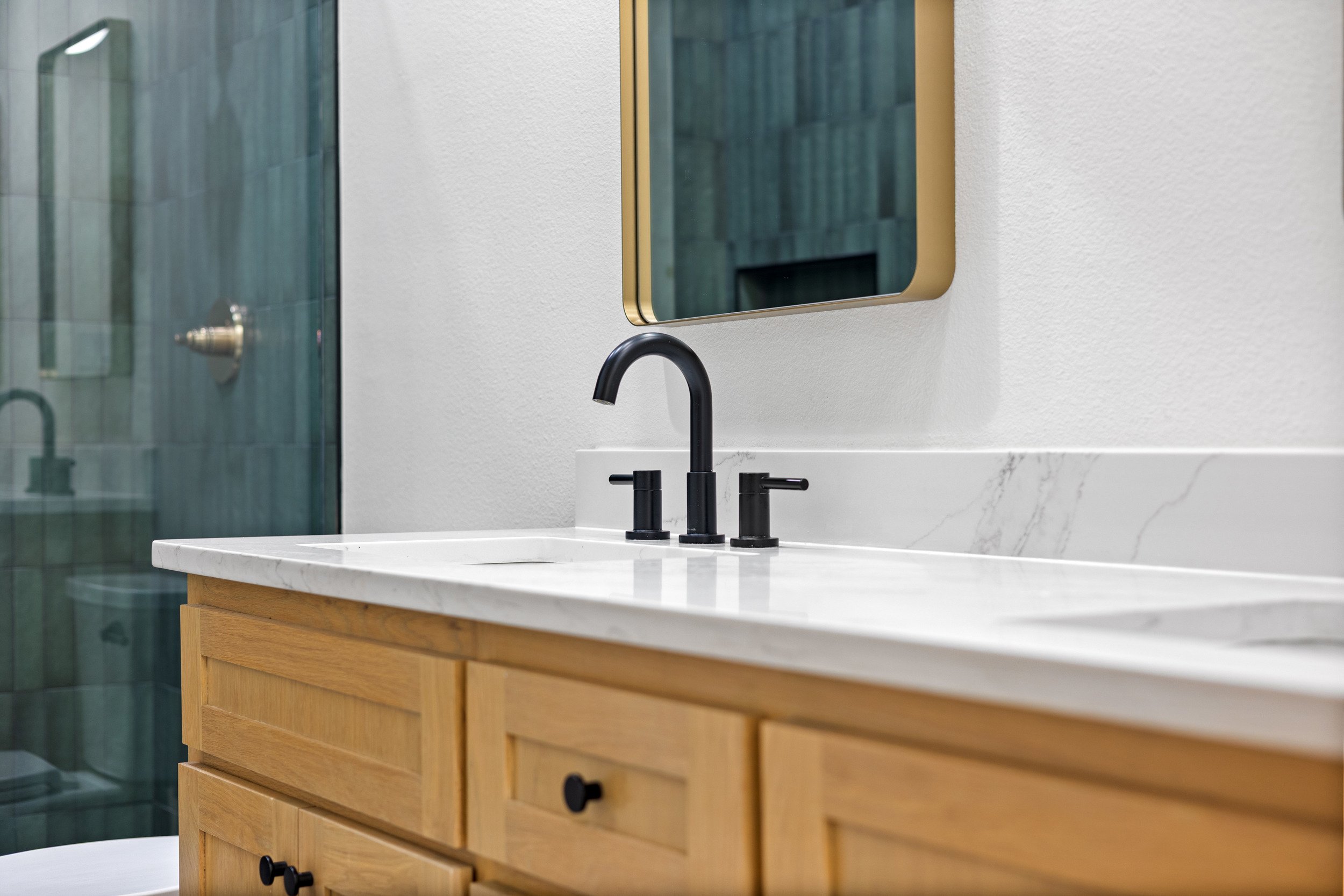 Bathroom vanity with a white quartz countertop, a black faucet, a mirror with a gold frame, wooden cabinet, and a glass shower enclosure in the background.