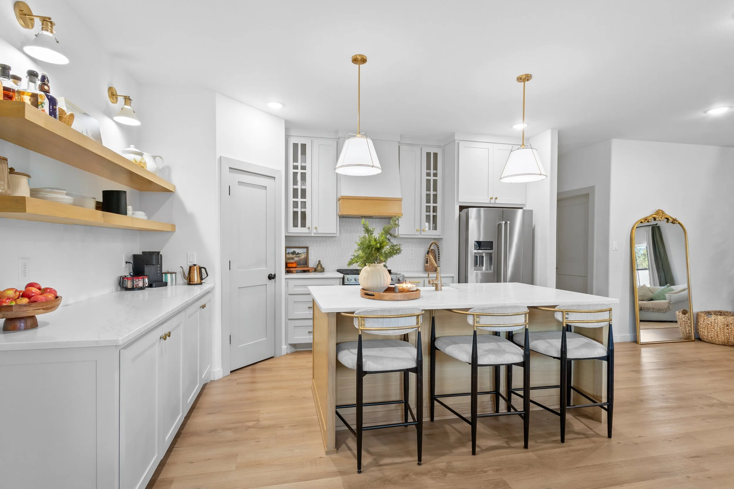 Modern kitchen with white cabinets, a large island with a white countertop, three barstools with light cushions, two pendant lights, a stainless steel refrigerator, open wooden shelves with dishes, and a mirror leaning against the wall near a woven b