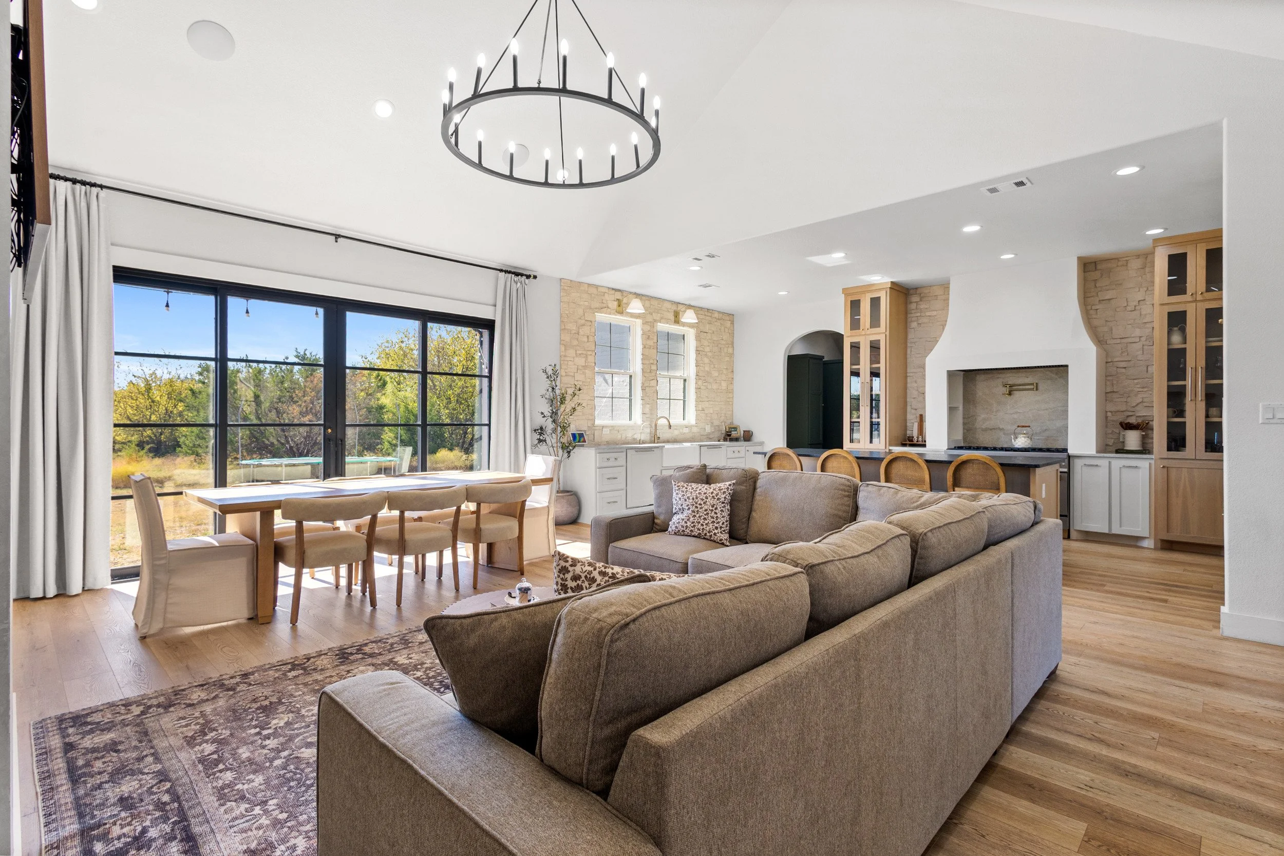 Open living room and kitchen with large glass sliding doors, beige sectional sofa, wooden dining table with chairs, and a modern chandelier.