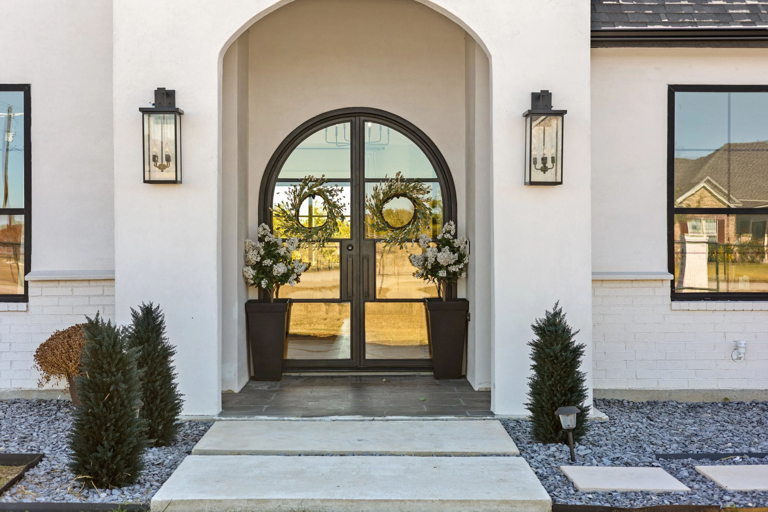 Front entrance of a modern house with a glass door, adorned with floral wreaths, flanked by potted white flower arrangements. The house has white walls, black lantern-style wall sconces, and surrounding landscaping with small shrubs, gravel, and step