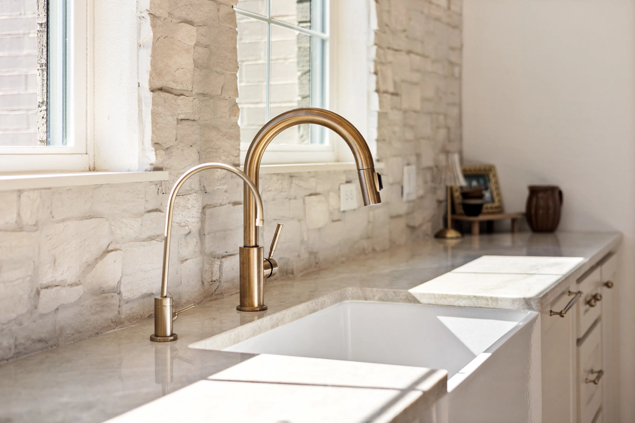 Kitchen sink with two brass faucets and a window above, featuring a stone backsplash and beige cabinetry, with decorative items on the counter.