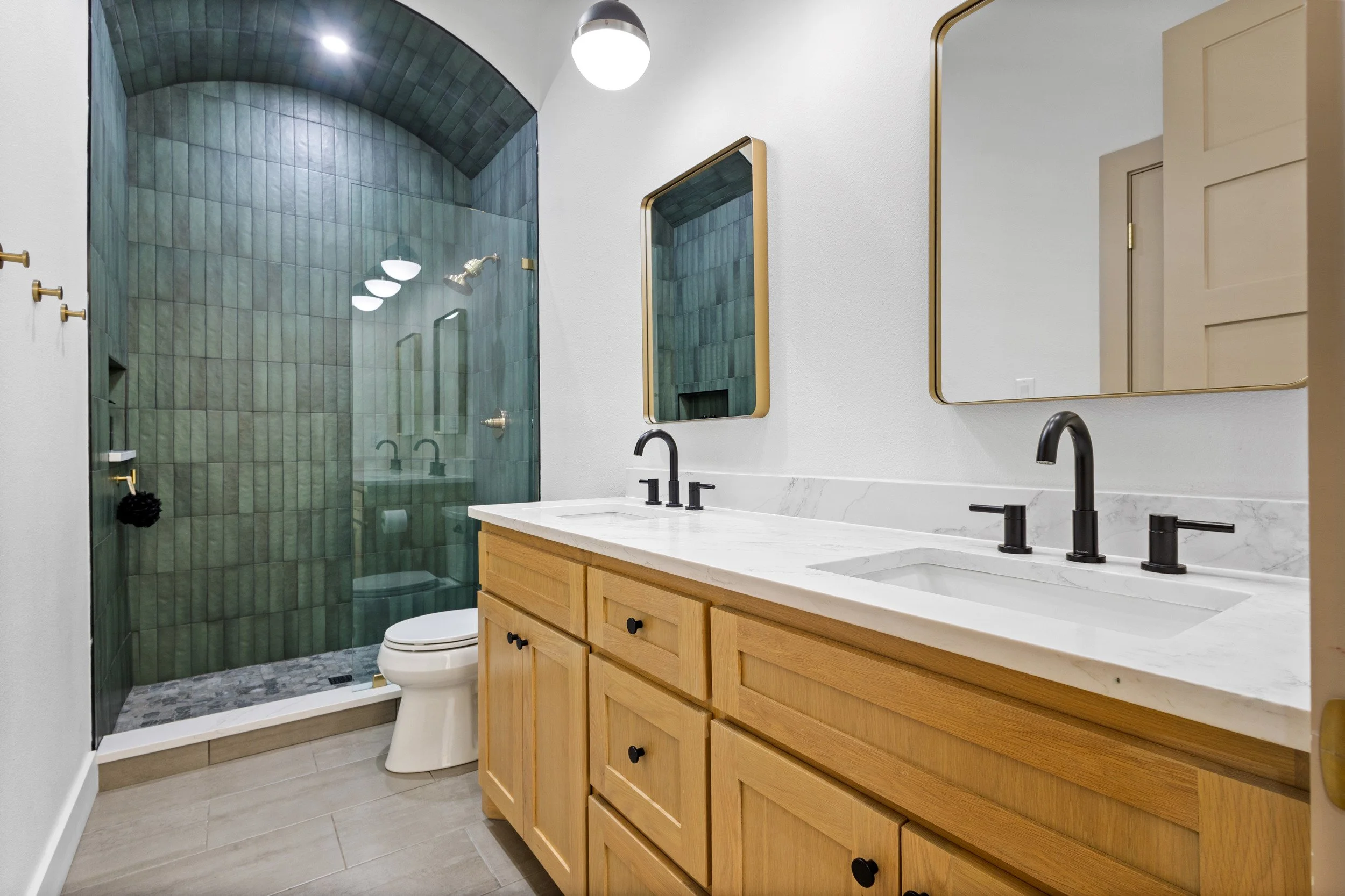 Modern bathroom with dual sinks, wooden vanity, large rectangular mirrors, and a green-tiled walk-in shower with a curved ceiling.
