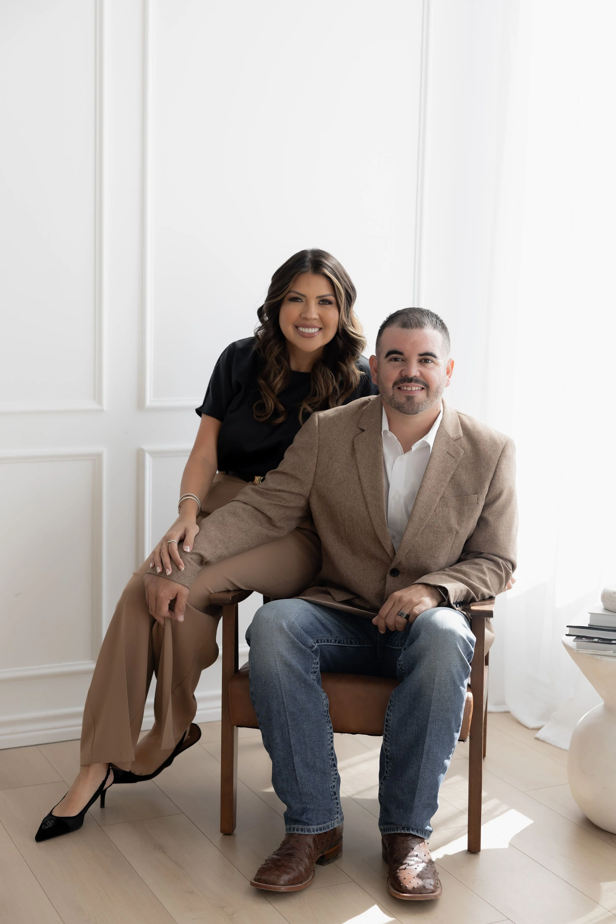 A woman and a man sitting in a bright, white room with paneled walls. The woman is standing slightly behind and to the left of the man, smiling, with her hand resting on his shoulder. She has dark, wavy hair, and is dressed in black and beige professional attire. The man is sitting on a wooden chair, wearing a tan blazer, white shirt, jeans, and brown boots, smiling at the camera.