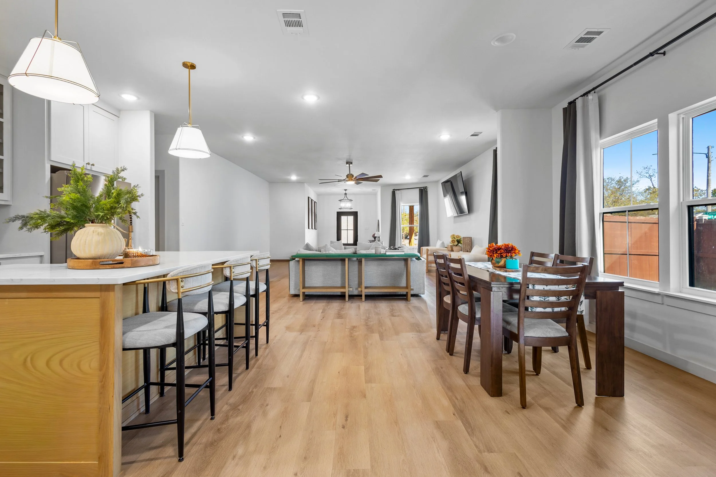 Open-concept living and dining area with hardwood floors, white walls, large windows, and modern furnishings including a kitchen island, dining table with chairs, and a sofa with a television.