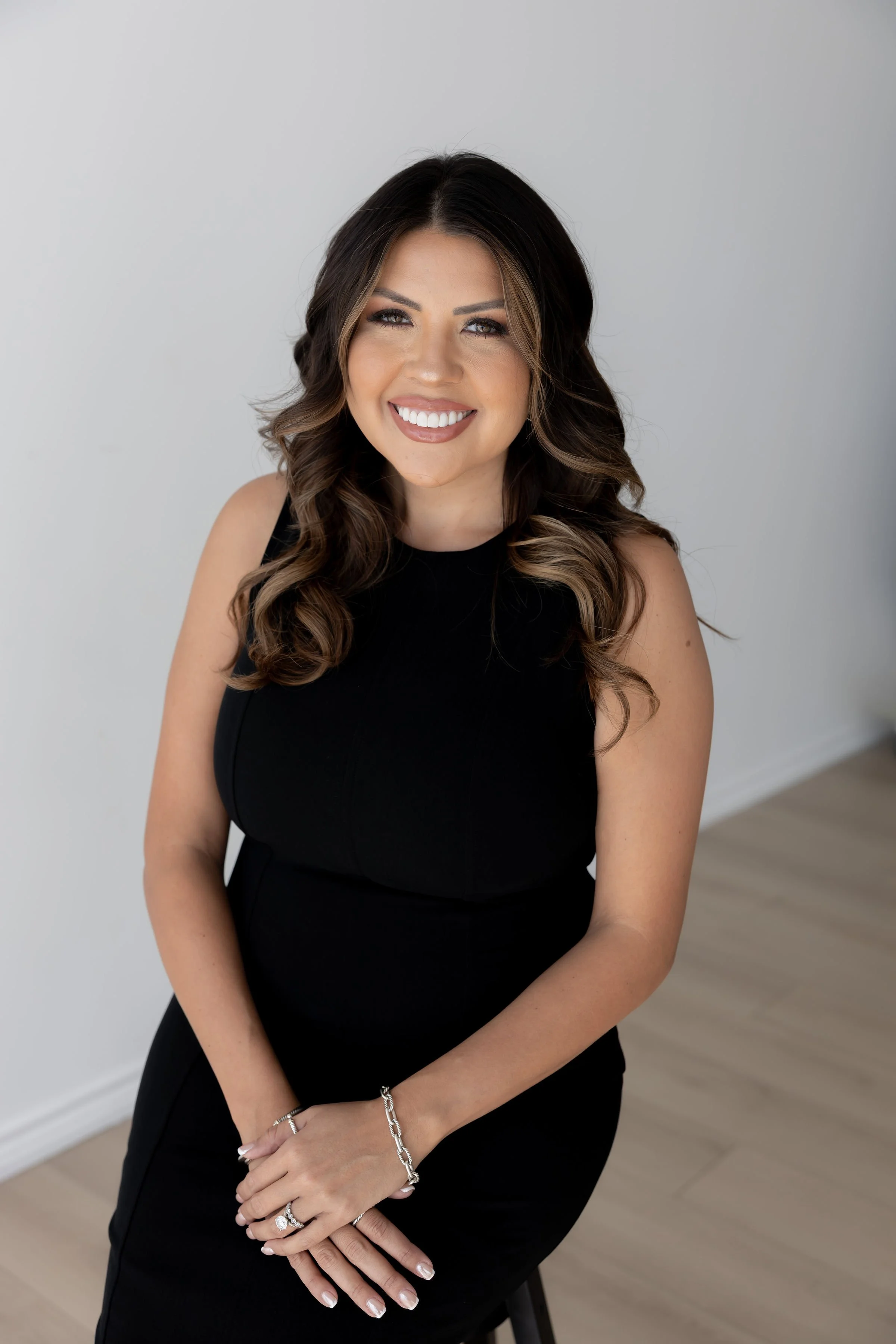 A woman with long, wavy brown hair, wearing a sleeveless black dress and silver jewelry, smiling at the camera in a bright room with a white wall and light wooden floor.