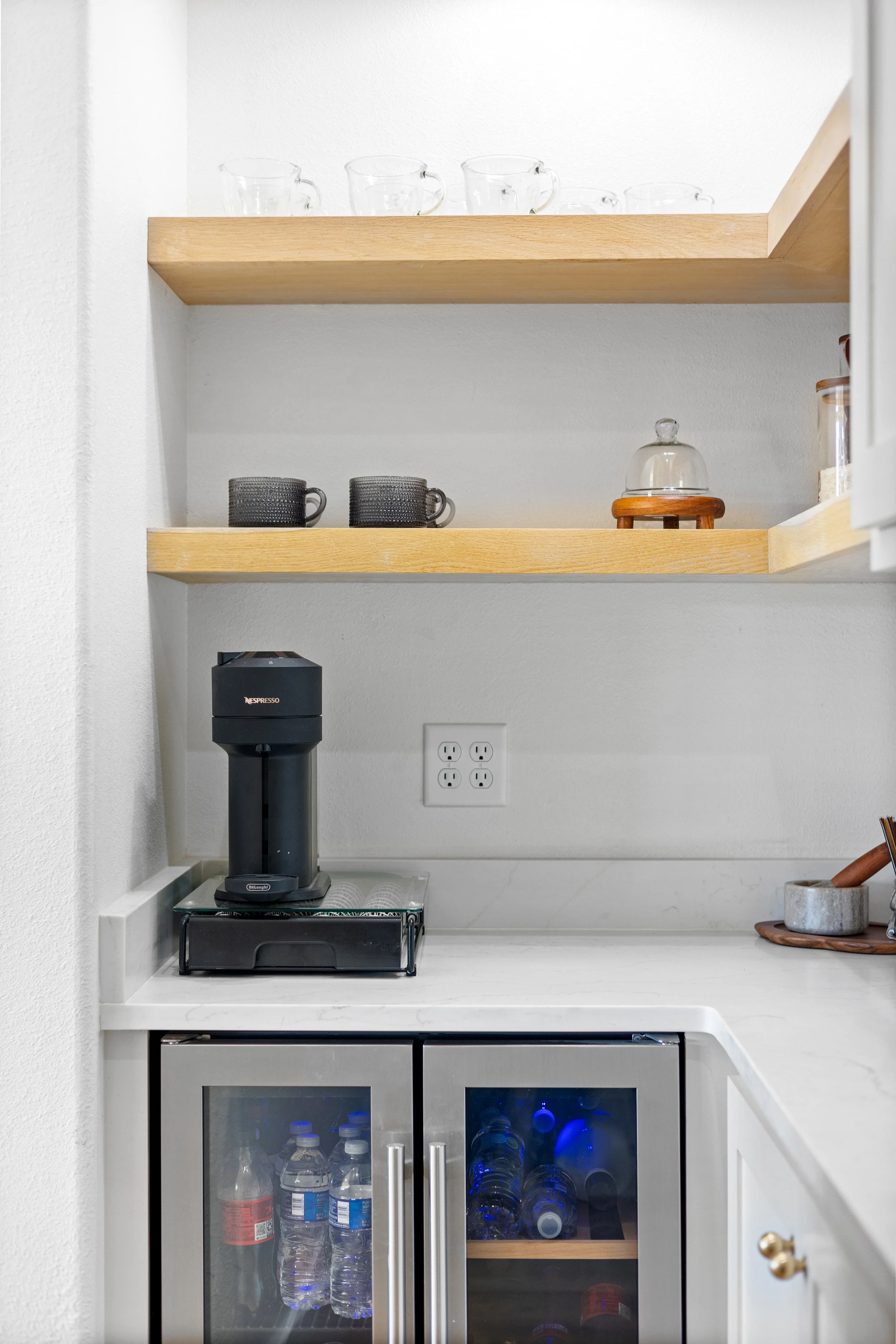 Kitchen corner with open wooden shelves holding glass cups, black and gray mugs, and a ceramic cloche. Below the shelves, a coffee machine is on the countertop next to a power outlet, with a mini fridge filled with bottled water underneath.