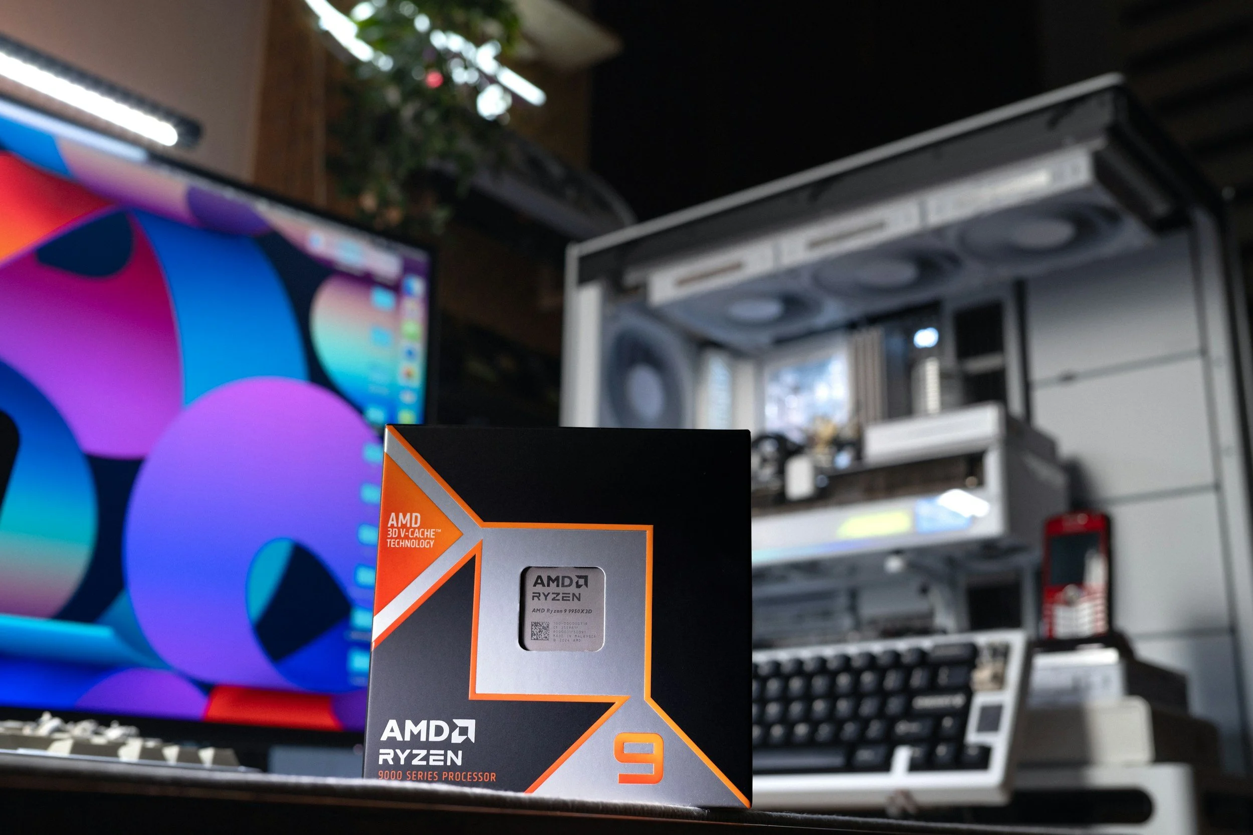 Box of AMD Ryzen 9 9000 series processor in front of a computer setup with a colorful monitor and a white computer case with fans.