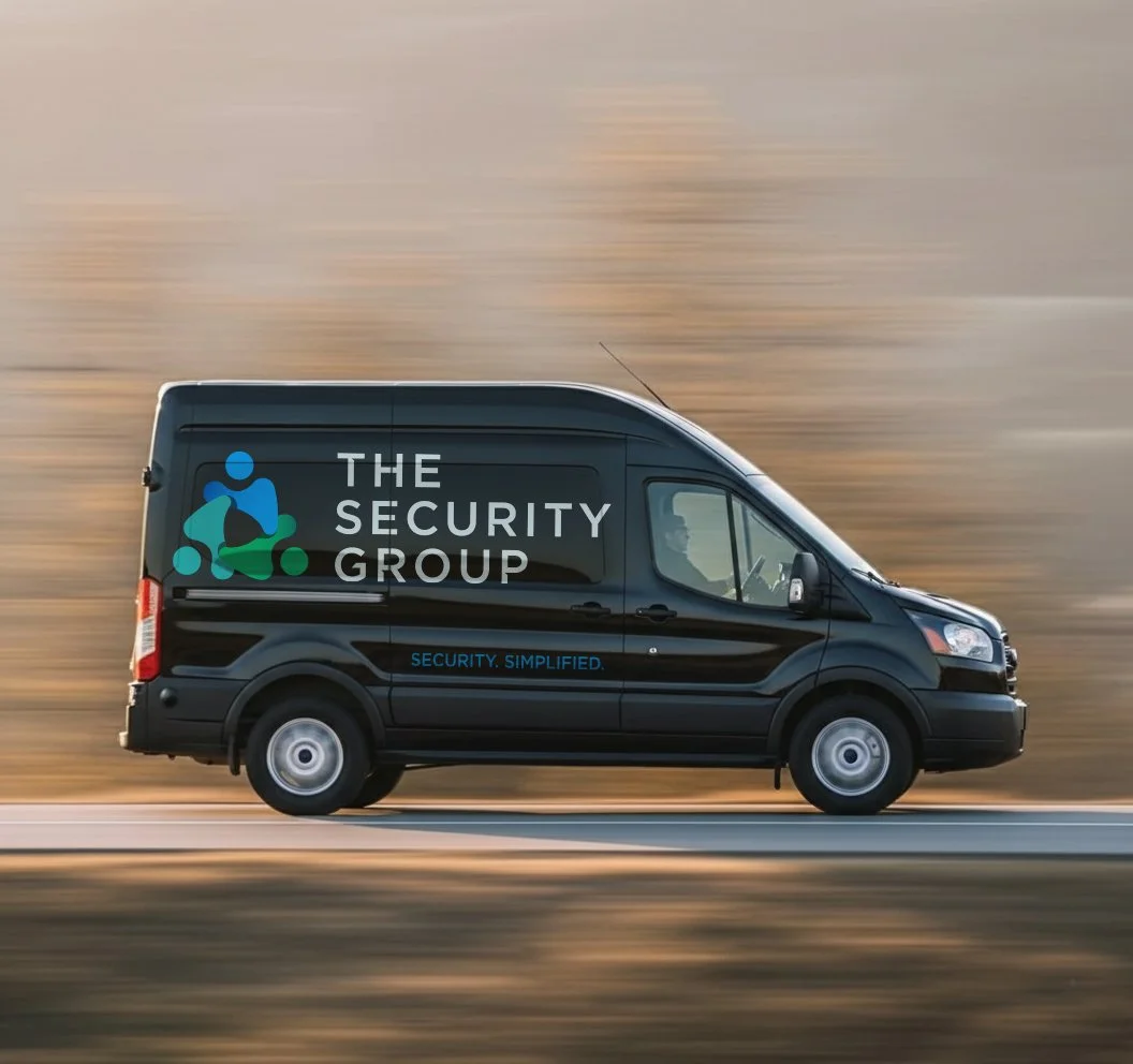 Black security vehicle with logo and text reading "The Security Group" and slogan "Security. Simplified." in motion on a highway with blurred background.