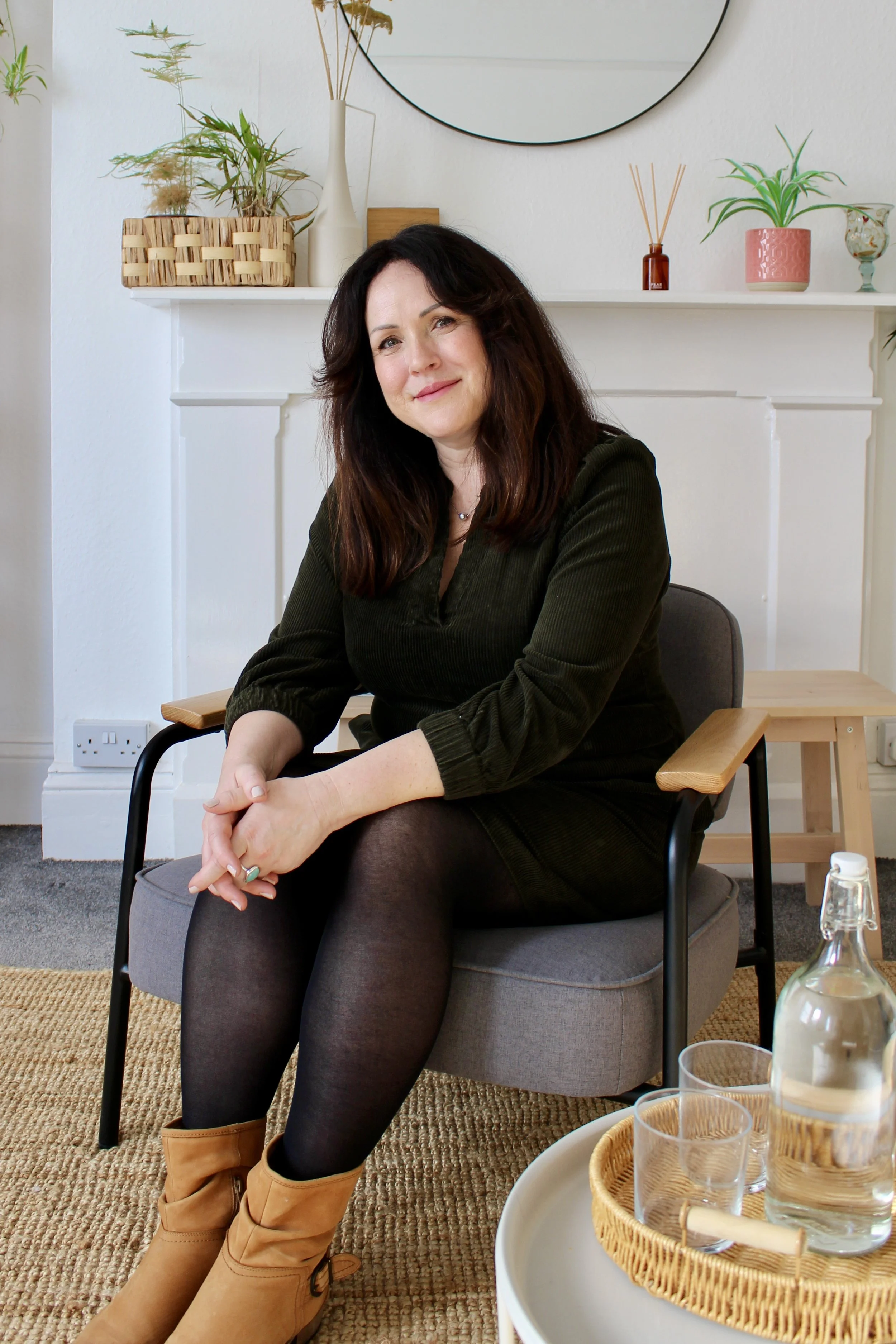 Ellen Baldwin wearing a dark green dress and tan boots, sitting on a gray armchair in her therapy room. There is a round mirror, potted plants, and decorative items on a white shelf behind her.
