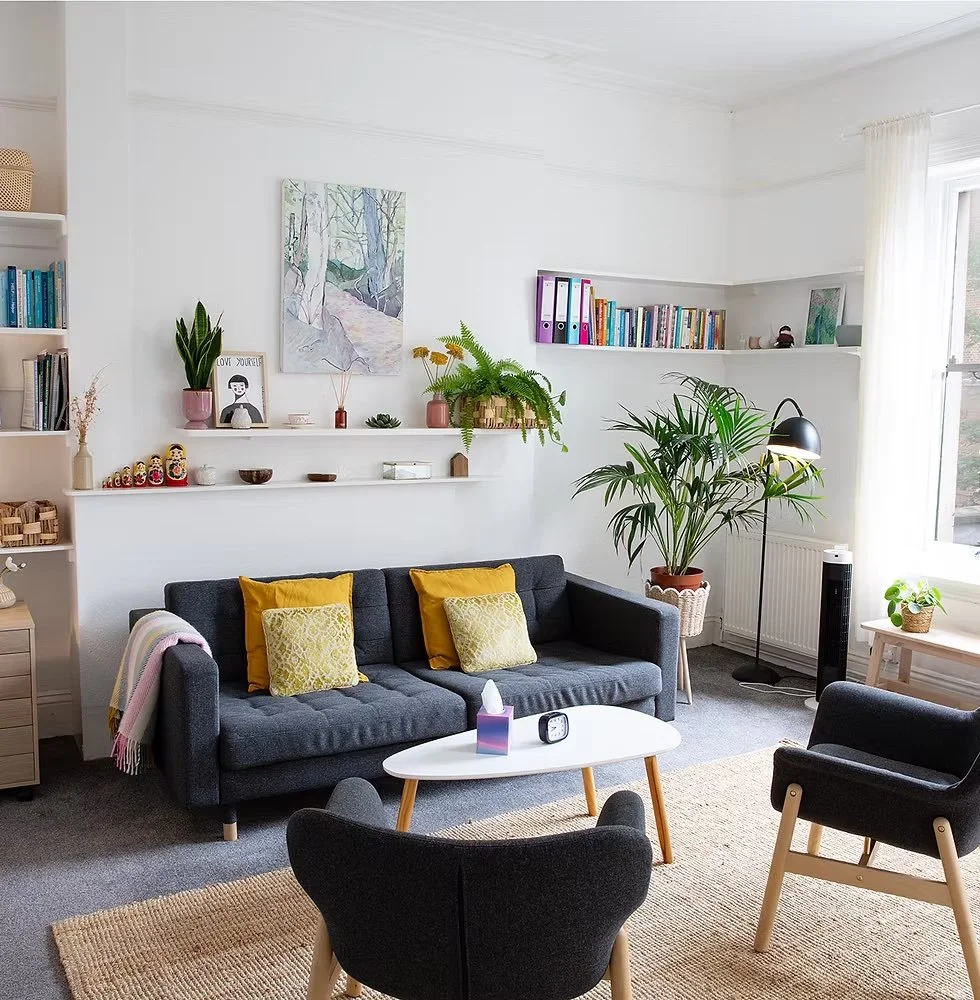 Therapy room with a dark gray sofa with yellow and light green pillows, a black armchair, a white oval coffee table, a tall green plant, and white walls with shelves holding books, plants, and decorative items. Natural light coming through a window.