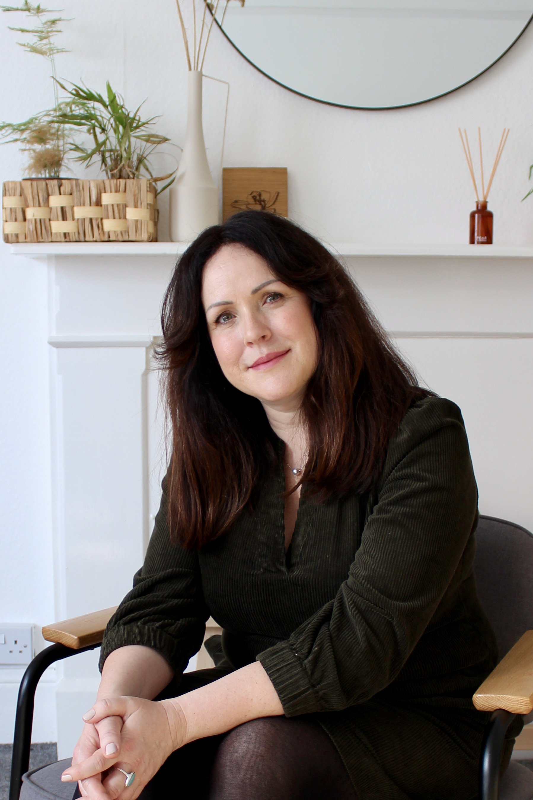 Ellen Baldwin sitting on a chair with wooden and black accents in her therapy room.