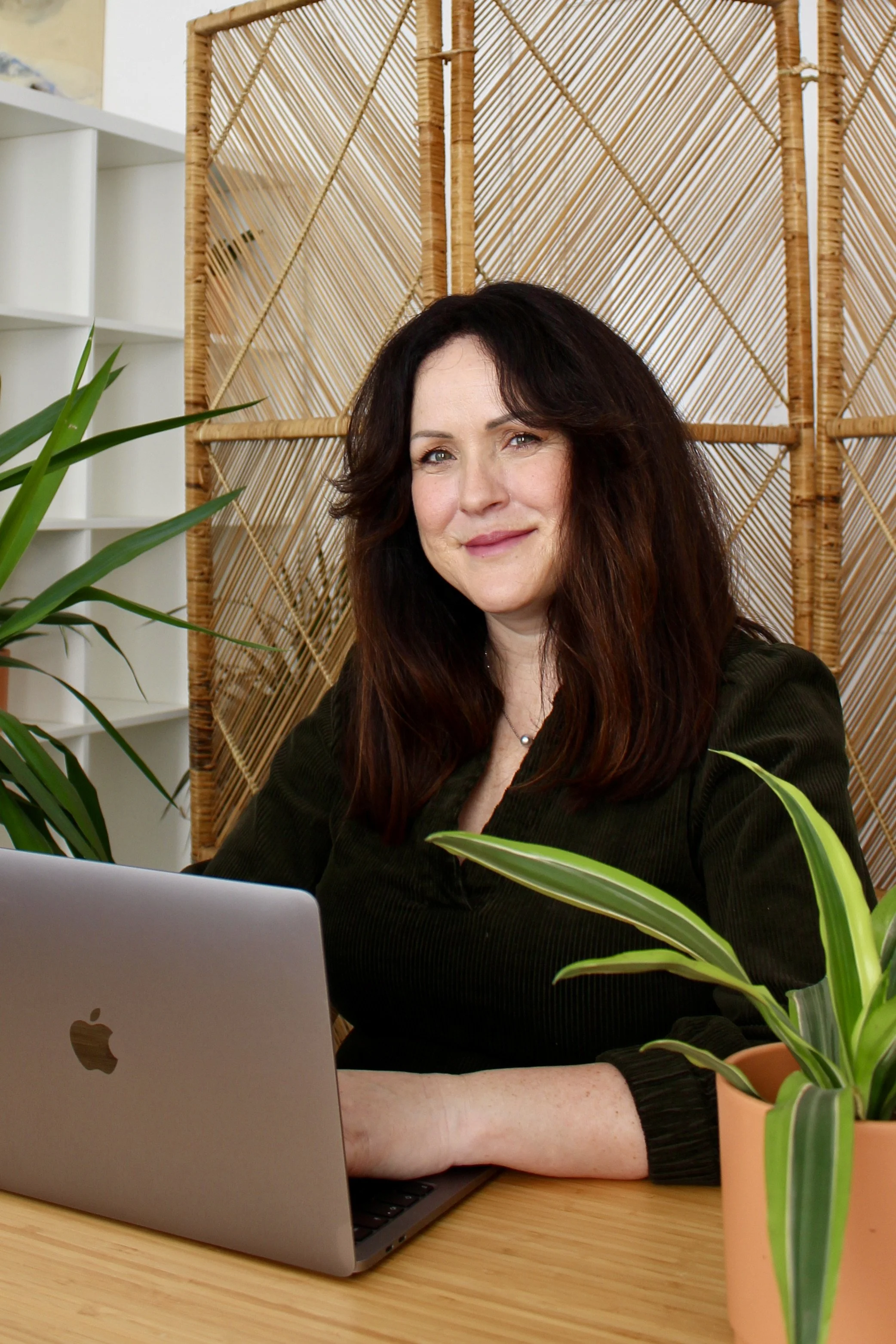 Ellen Baldwin smiling while sitting at a wooden table with a silver MacBook laptop in front of her, surrounded by green plants and a bamboo room divider behind her.