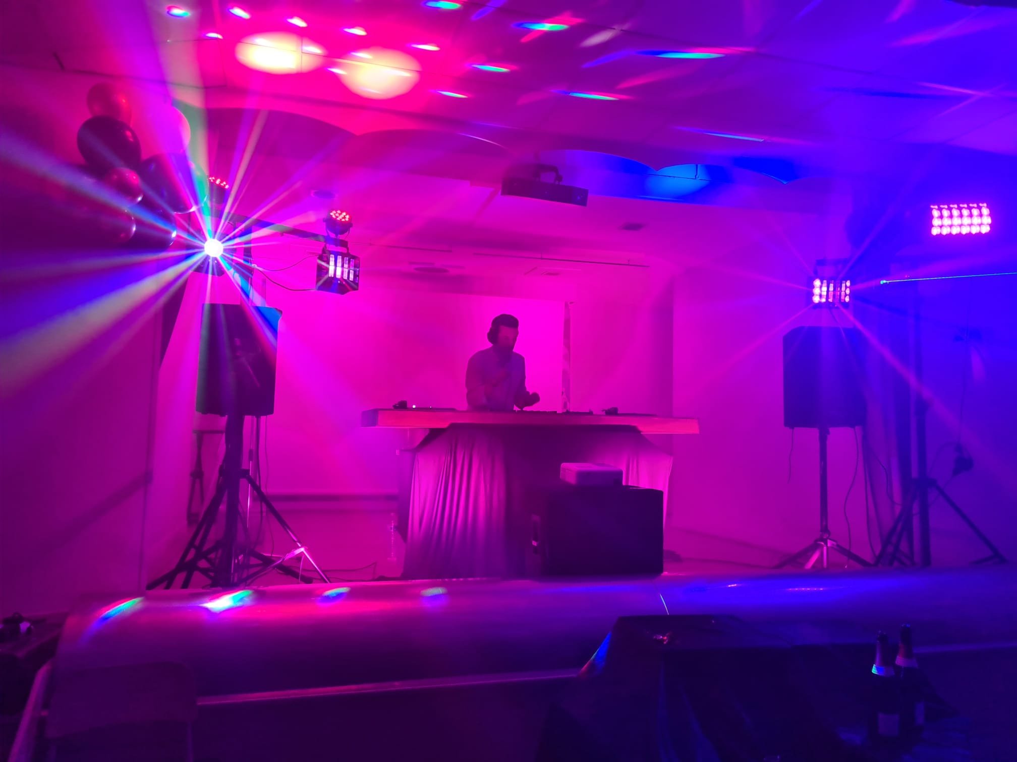 Un DJ joue dans une pièce avec un éclairage de discothèque violet et rose, entouré de lumières et de haut-parleurs.