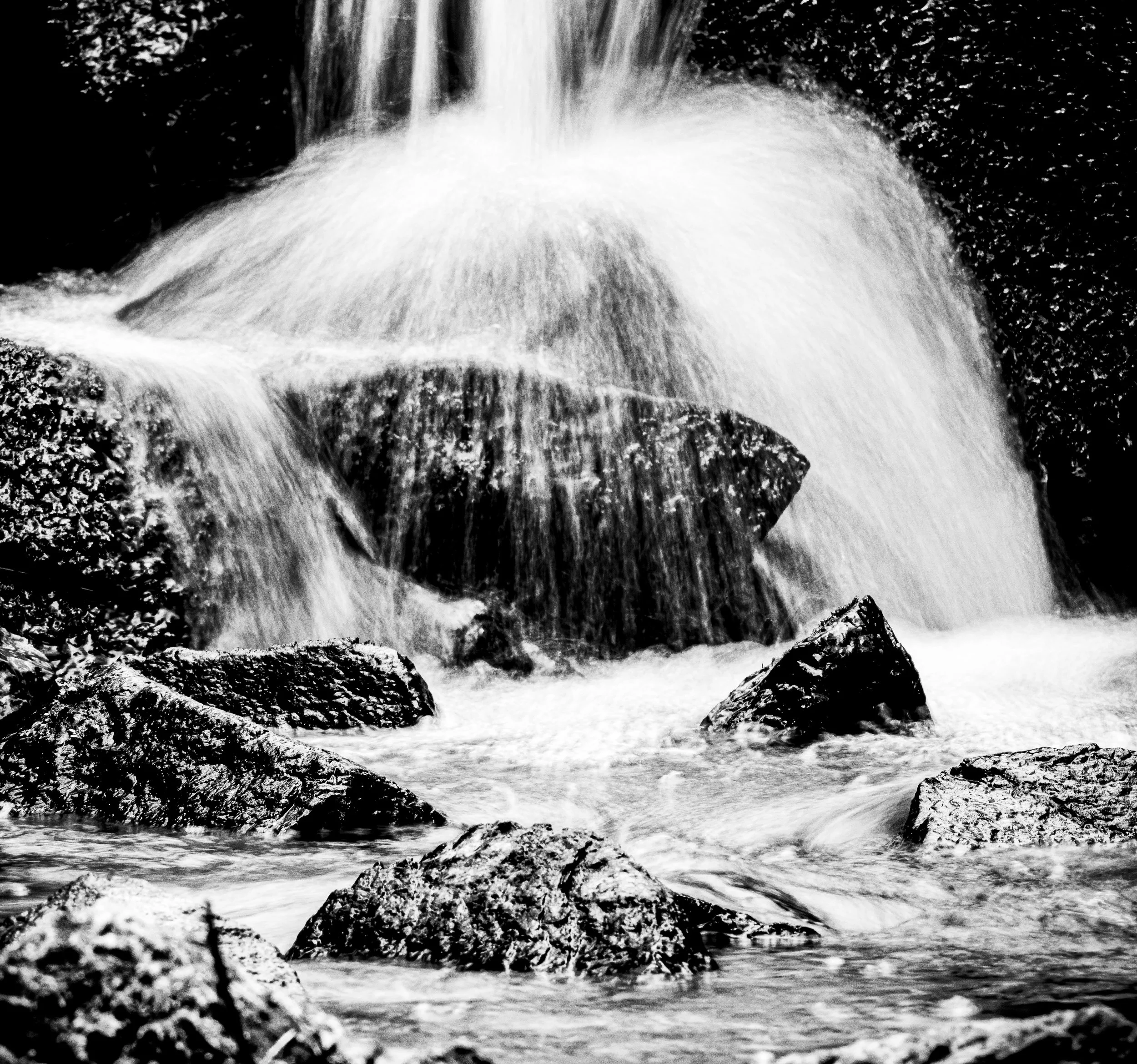 Black and white photograph of water flowing over rocks in a stream or small waterfall.